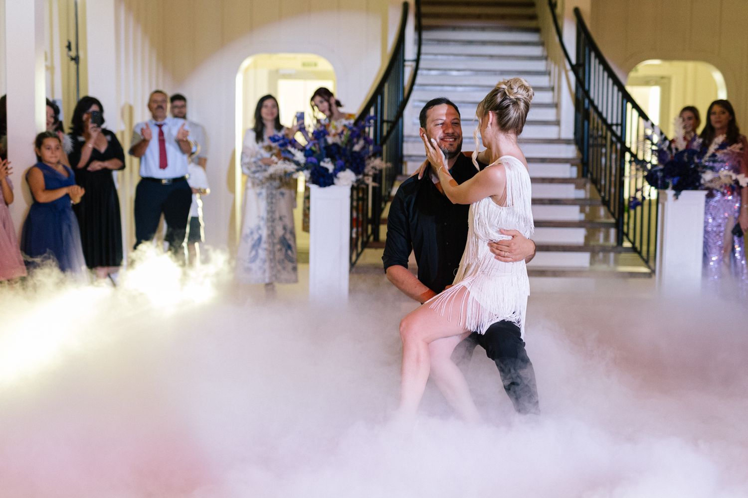 Wedding guests watch a couple dance in an elegant ballroom with soft lighting and ornate decor.