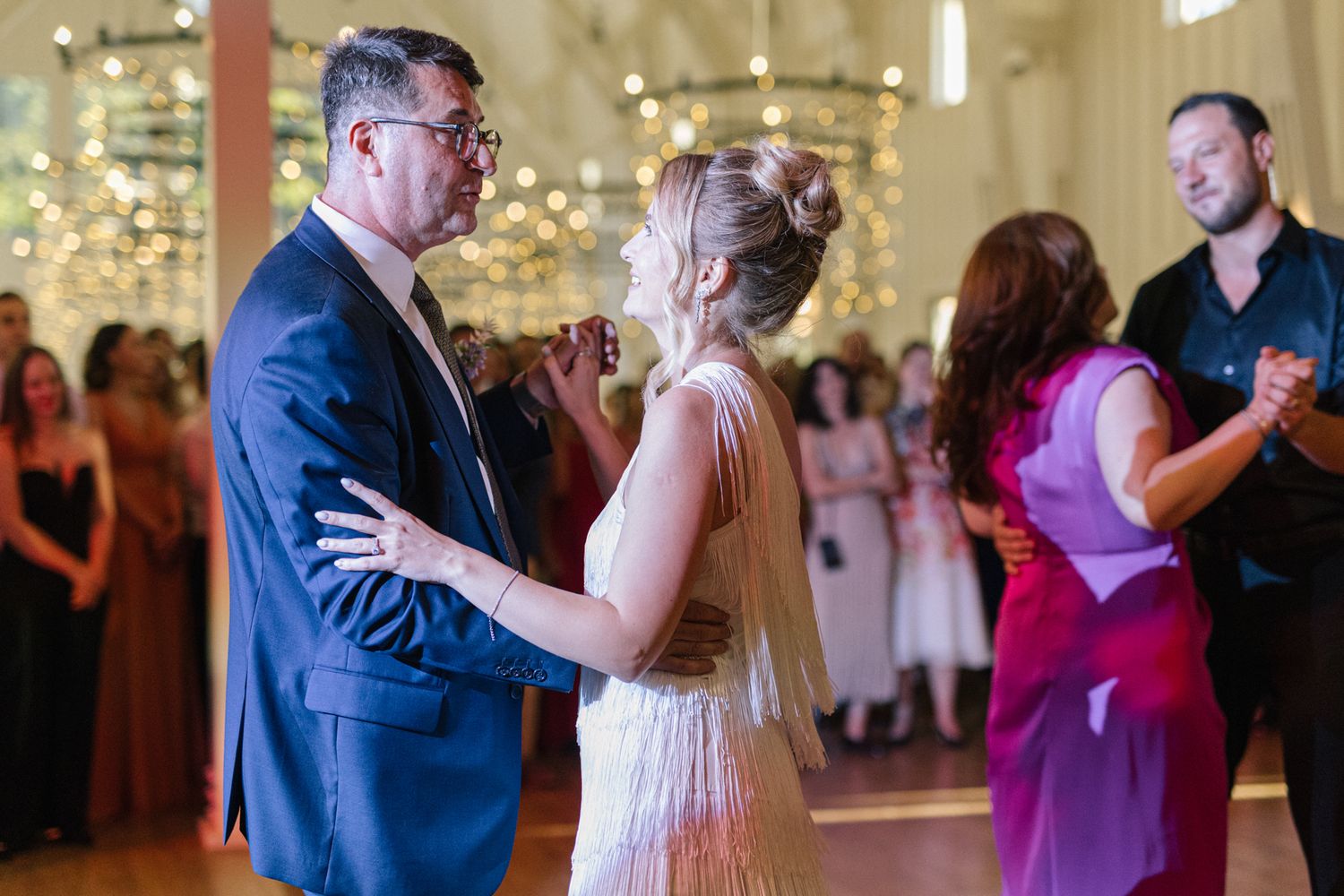Couples dancing at a formal wedding reception in an elegant white ballroom with crystal lighting.