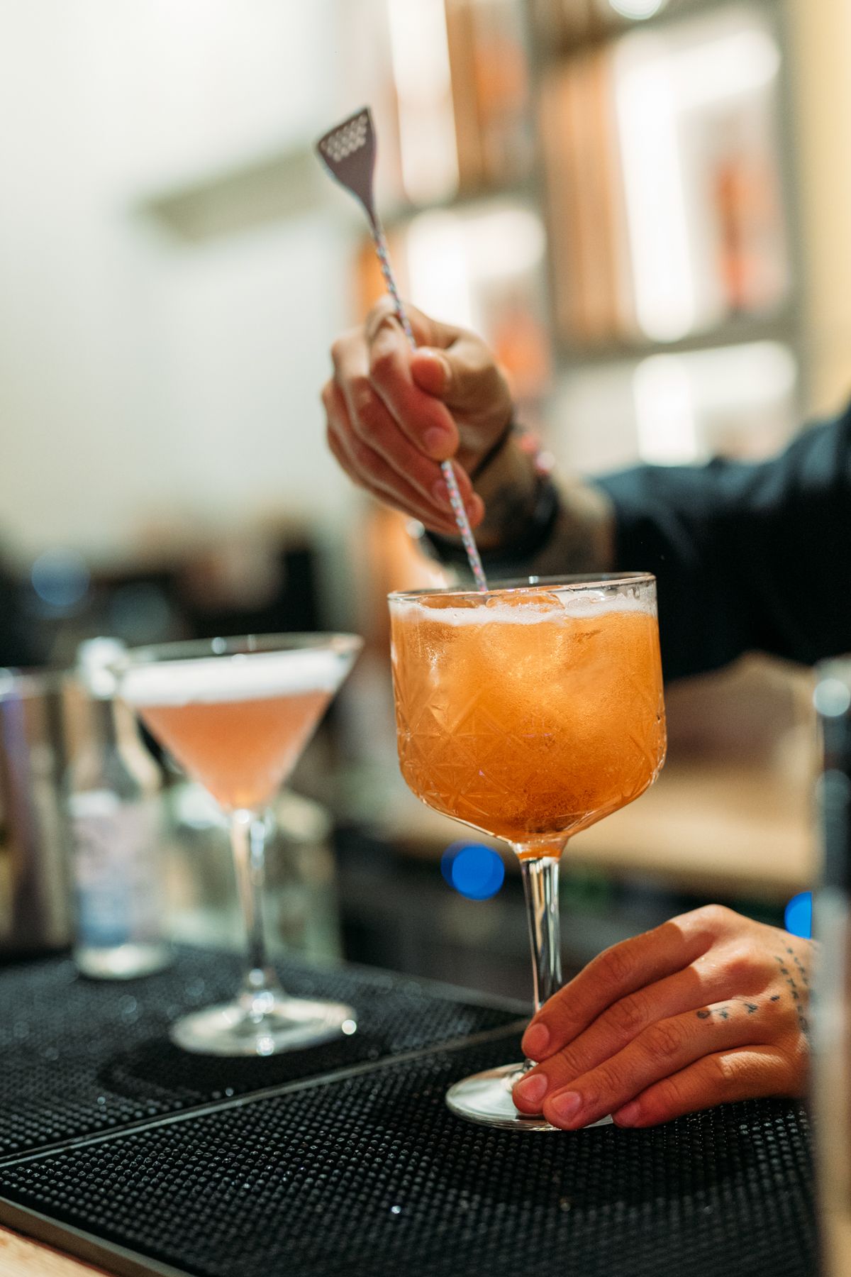 Orange cocktails being served on a black bar mat in elegant coupe glasses.