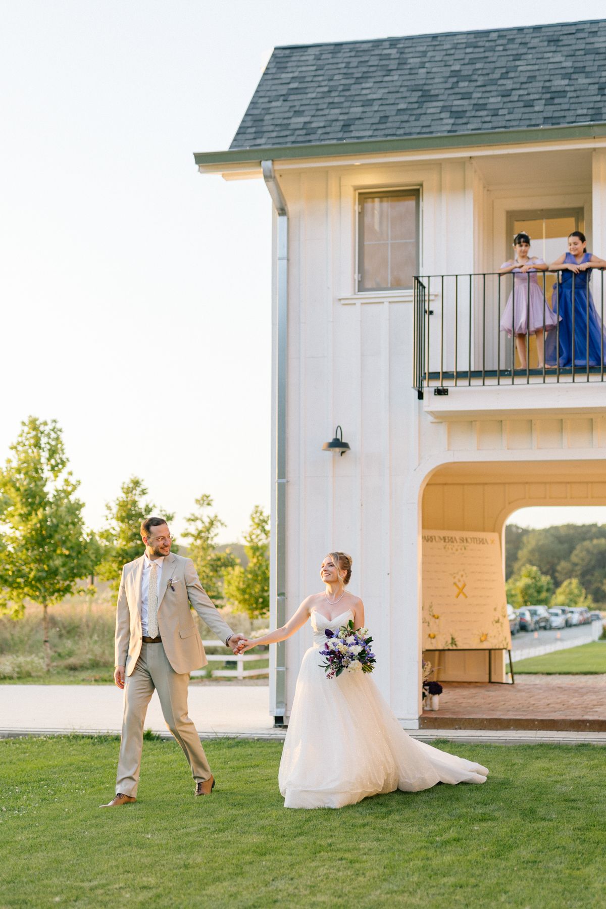 Wedding couple poses playfully on a grassy lawn beside a white farmhouse.