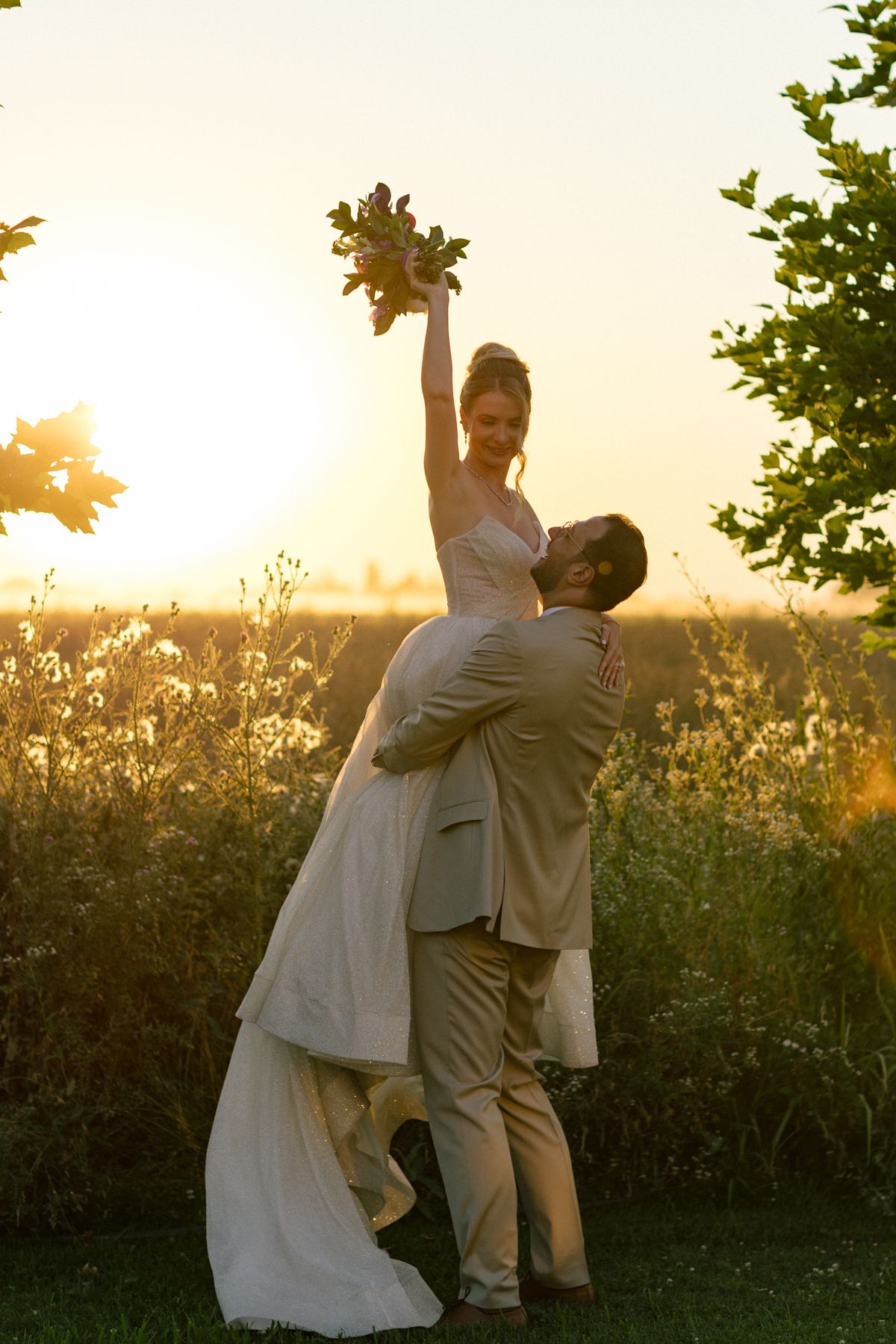 Wedding couple performs romantic lift pose against golden sunset backdrop.