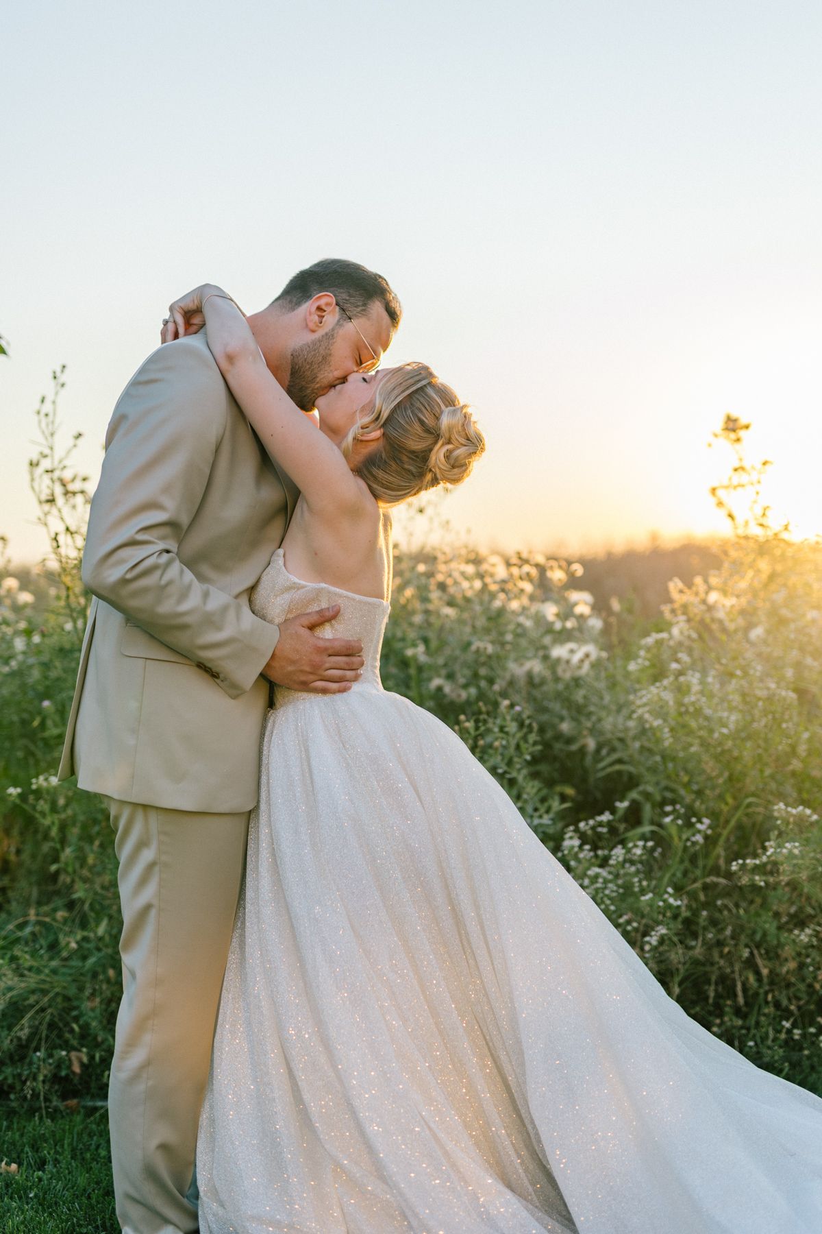 A couple in wedding attire embraces against a sunset backdrop with wildflowers.