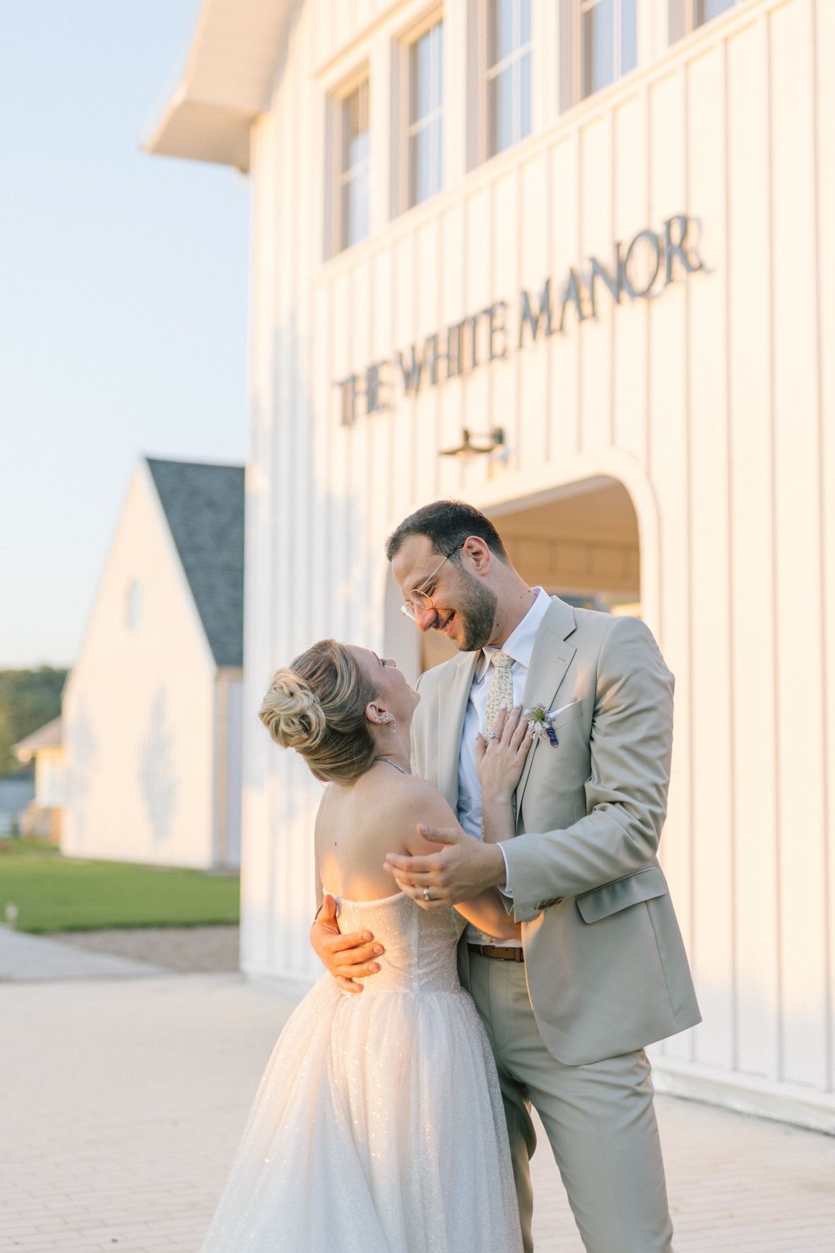 Newlyweds embrace in front of a white manor building at sunset.