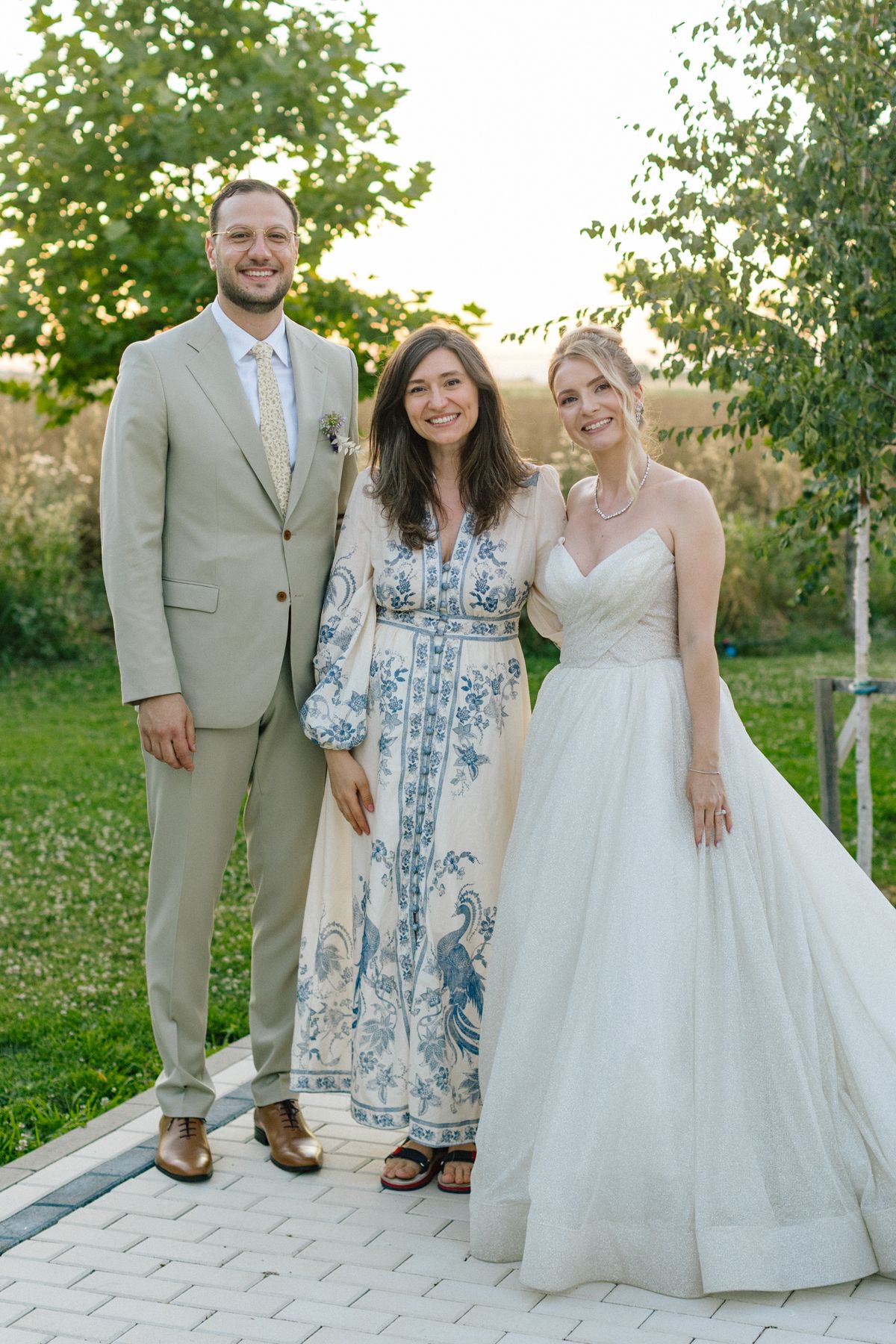 Wedding party poses outdoors in formal attire with tan suits and blue floral dress against natural backdrop.