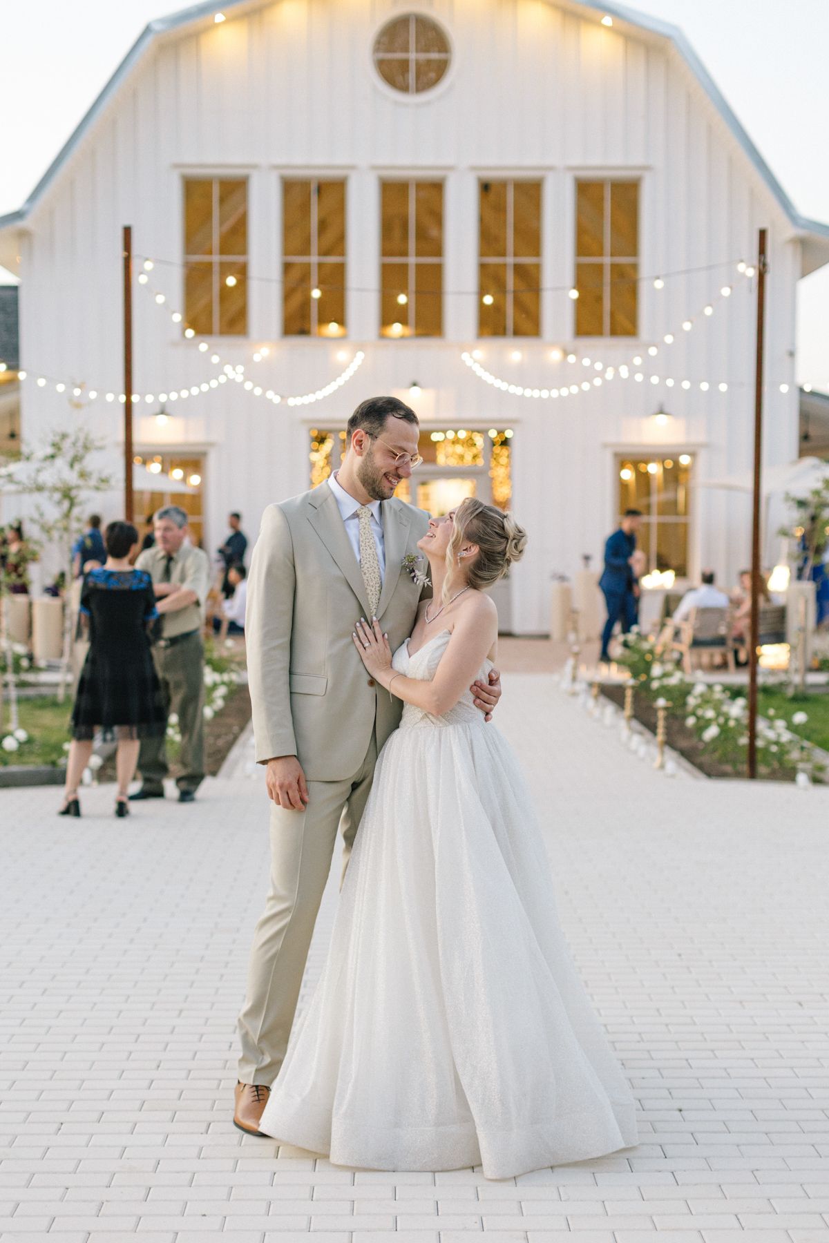 Couple celebrates at white barn wedding venue with string lights at dusk.