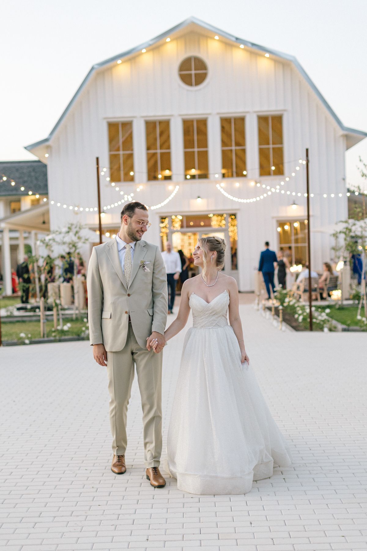 Couple walks together on stone path outside illuminated white barn wedding venue.