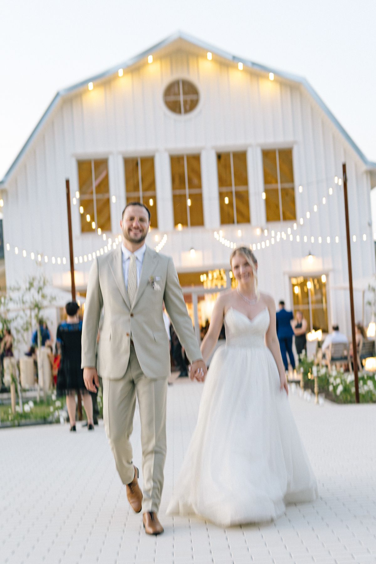 Wedding couple strolls along pathway near rustic white barn venue at sunset.