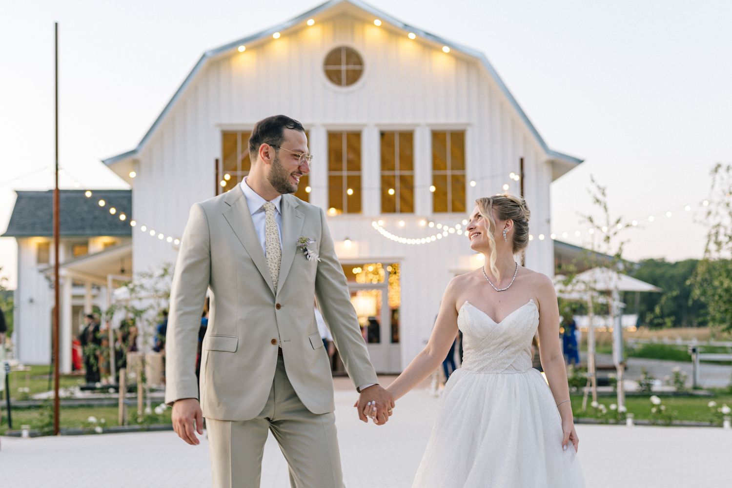 Couple walks hand in hand outside white barn wedding venue with string lights.