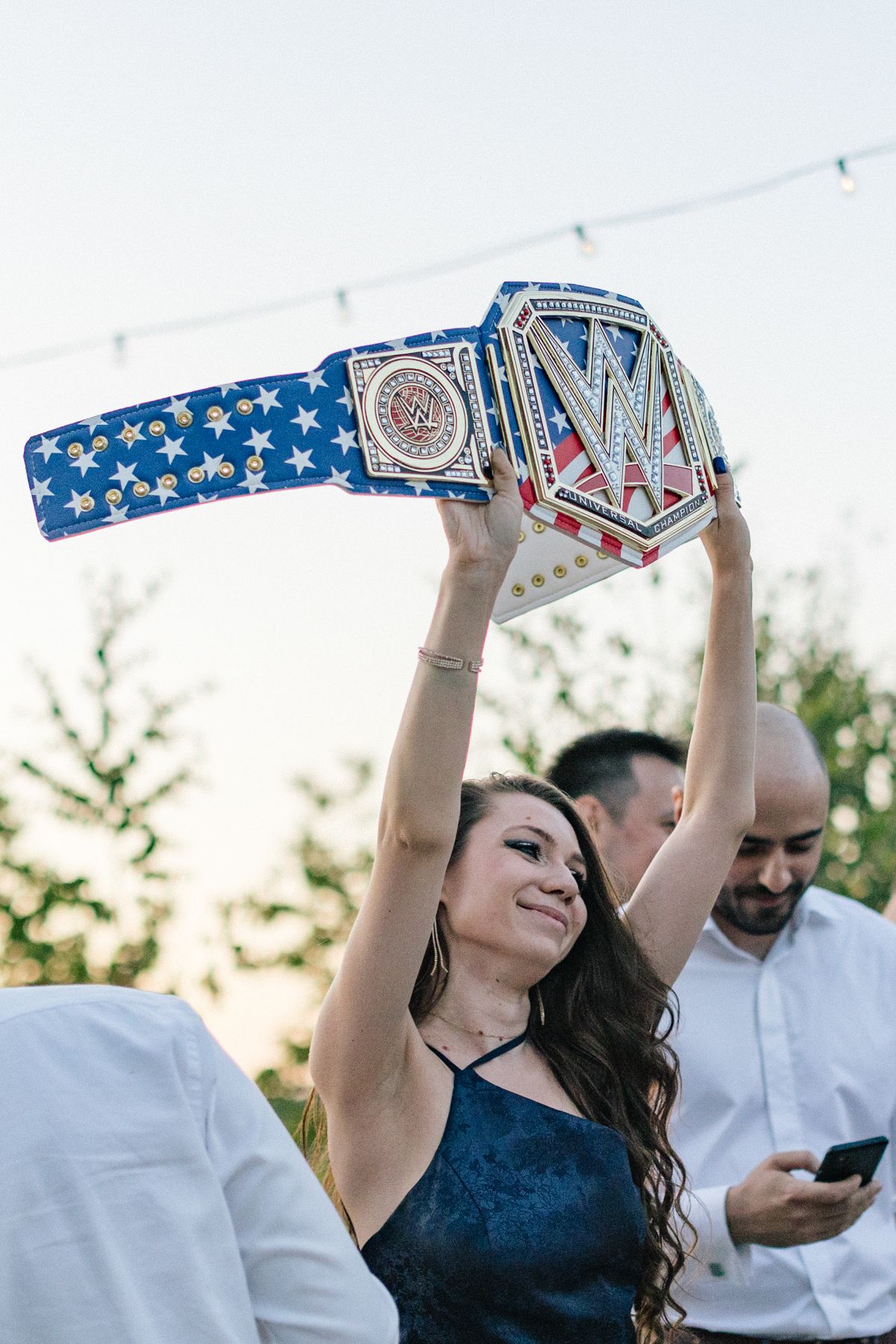 Guest holds up decorative blue and silver championship belt at outdoor wedding reception.