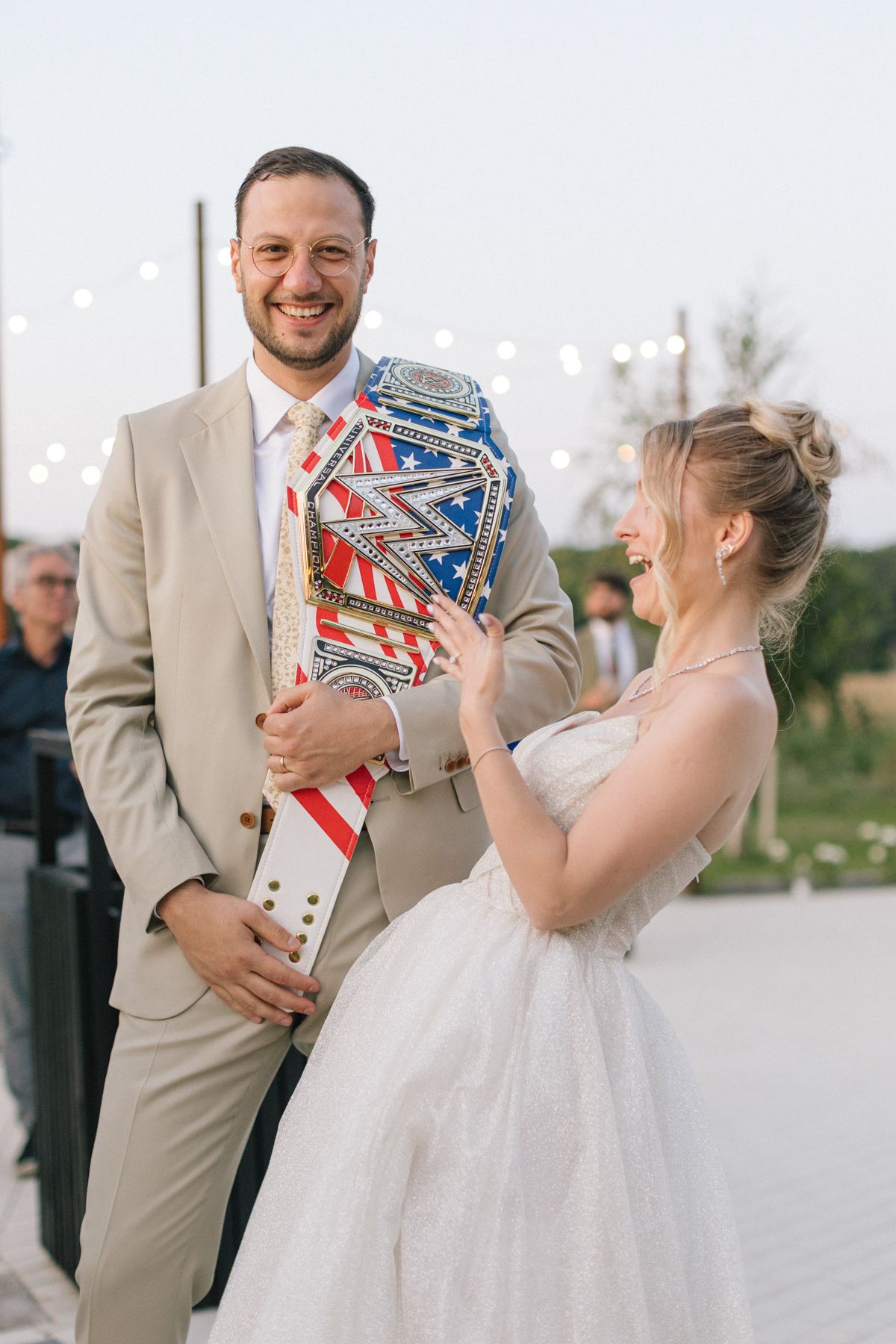 Couple shares playful moment with championship belt trophy at outdoor wedding reception.