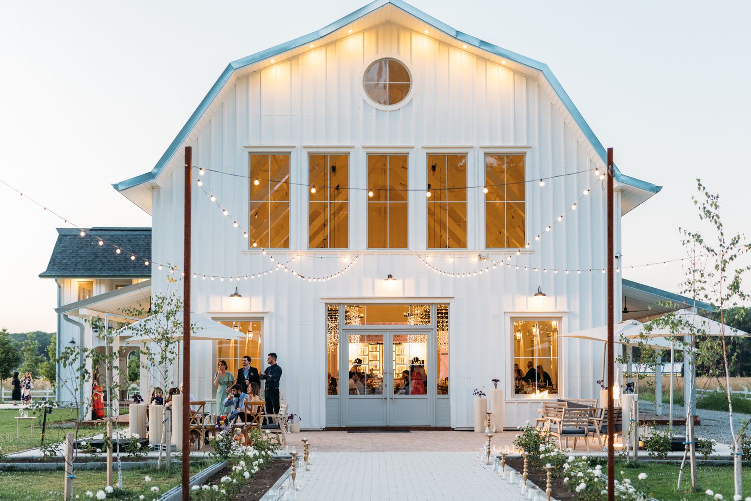 White barn wedding venue exterior with string lights and multiple windows at dusk.