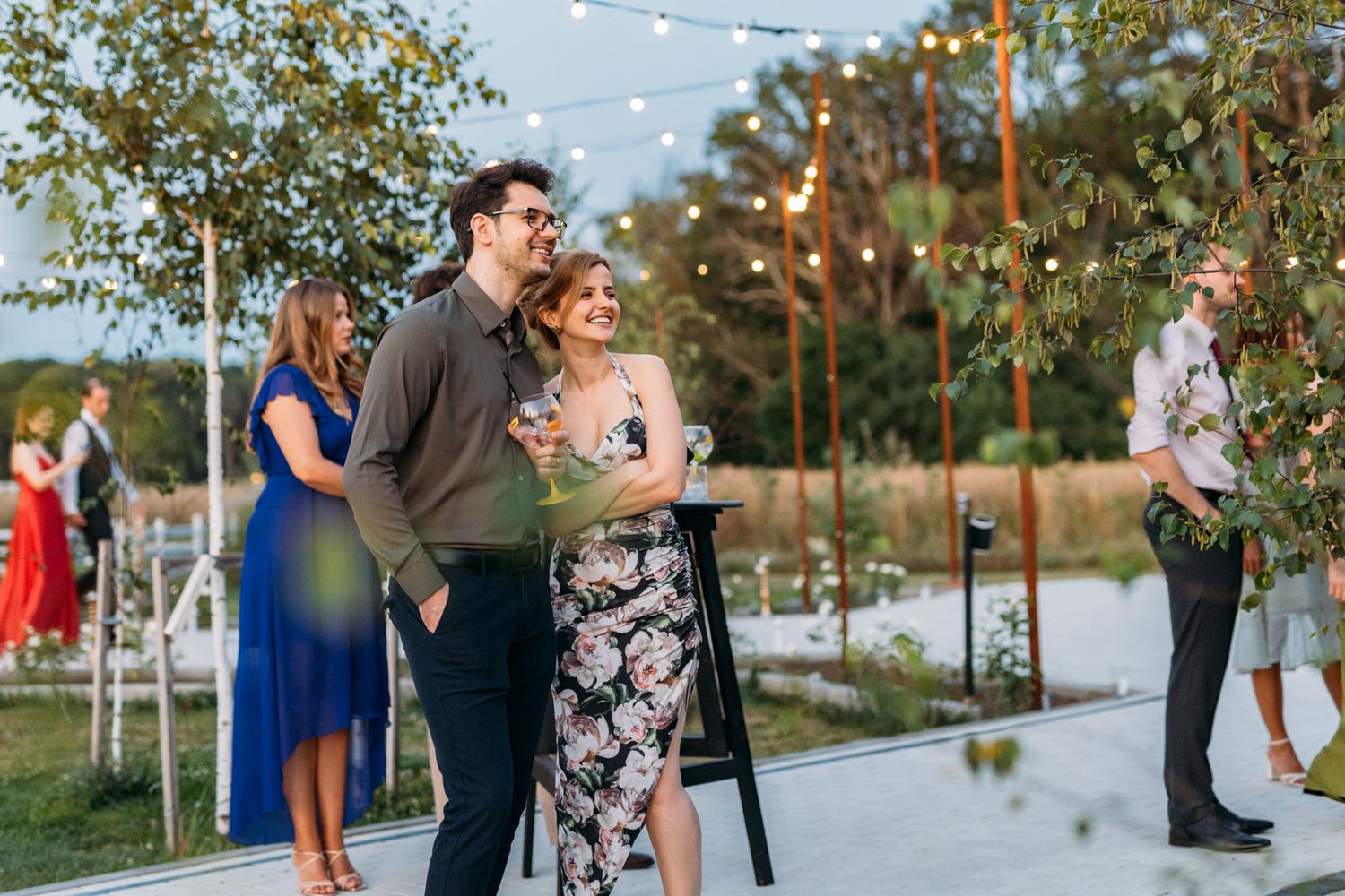 String lights illuminate an outdoor wedding reception as guests gather on a paved walkway.