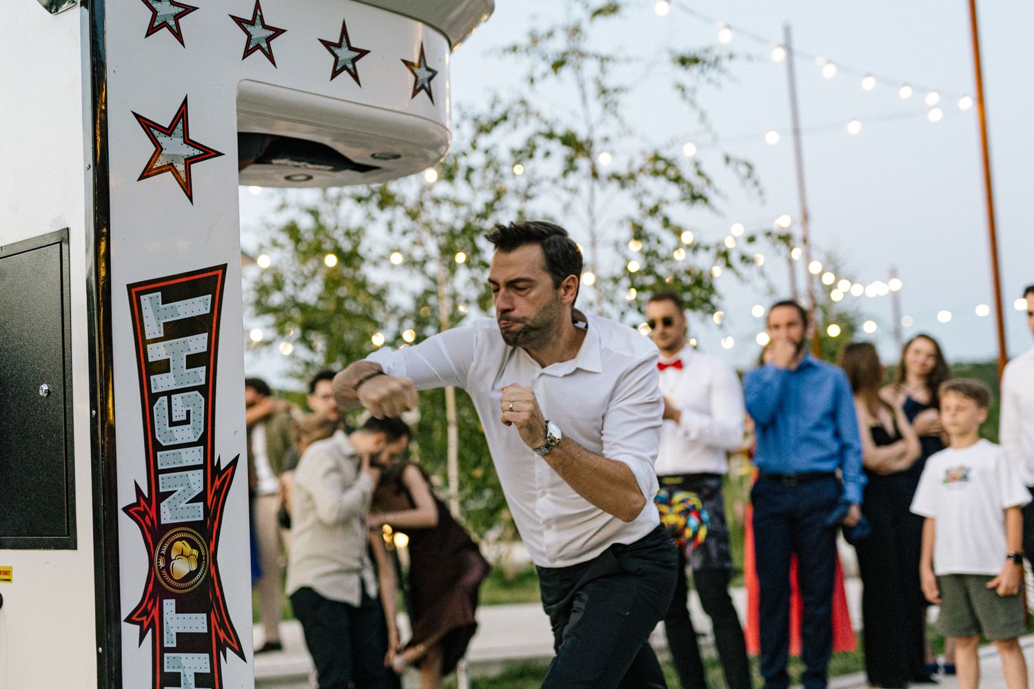 Wedding guests gather around an arcade-style punching game machine at an outdoor evening celebration.