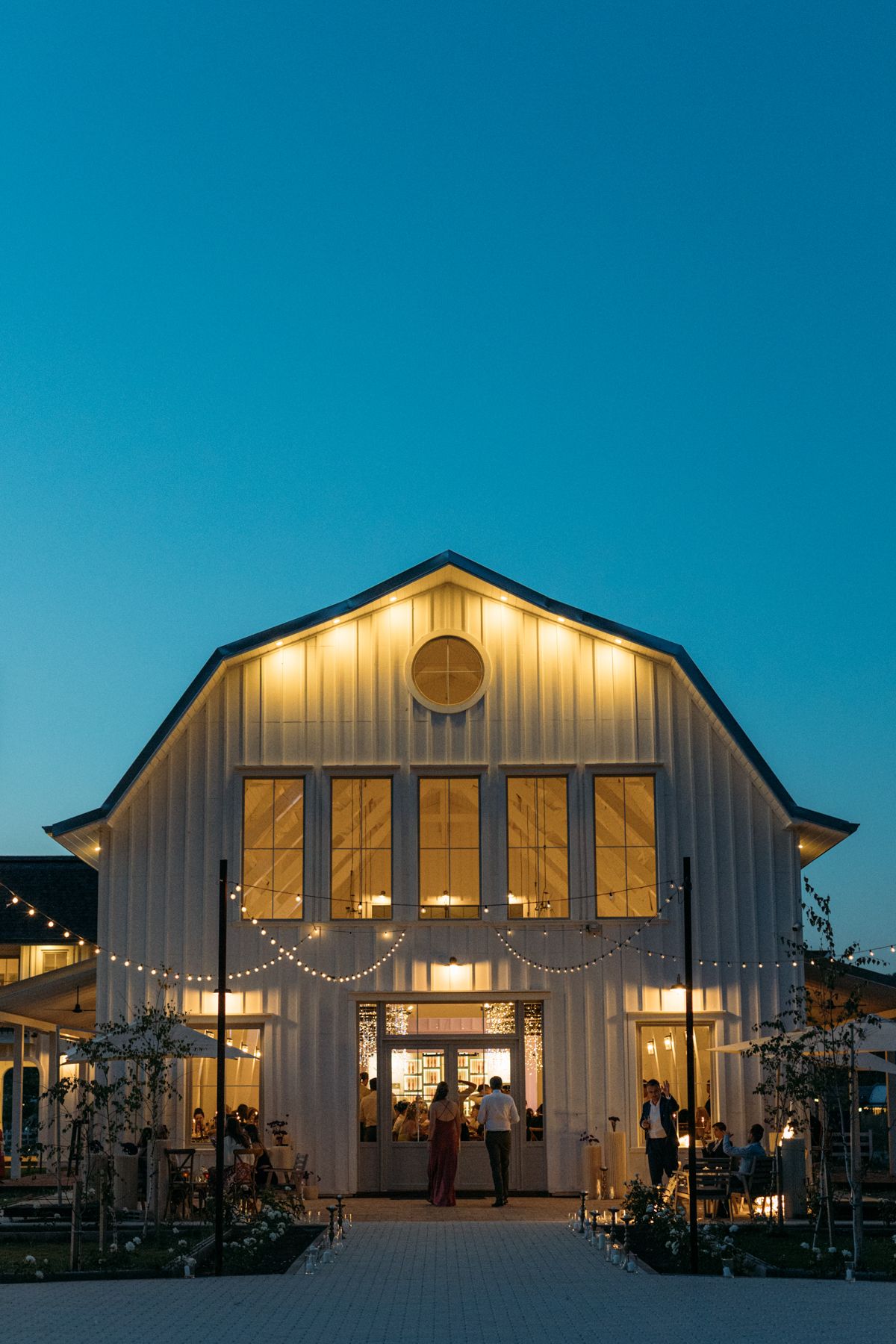 Modern white barn wedding venue illuminated at dusk with string lights and warm interior glow.