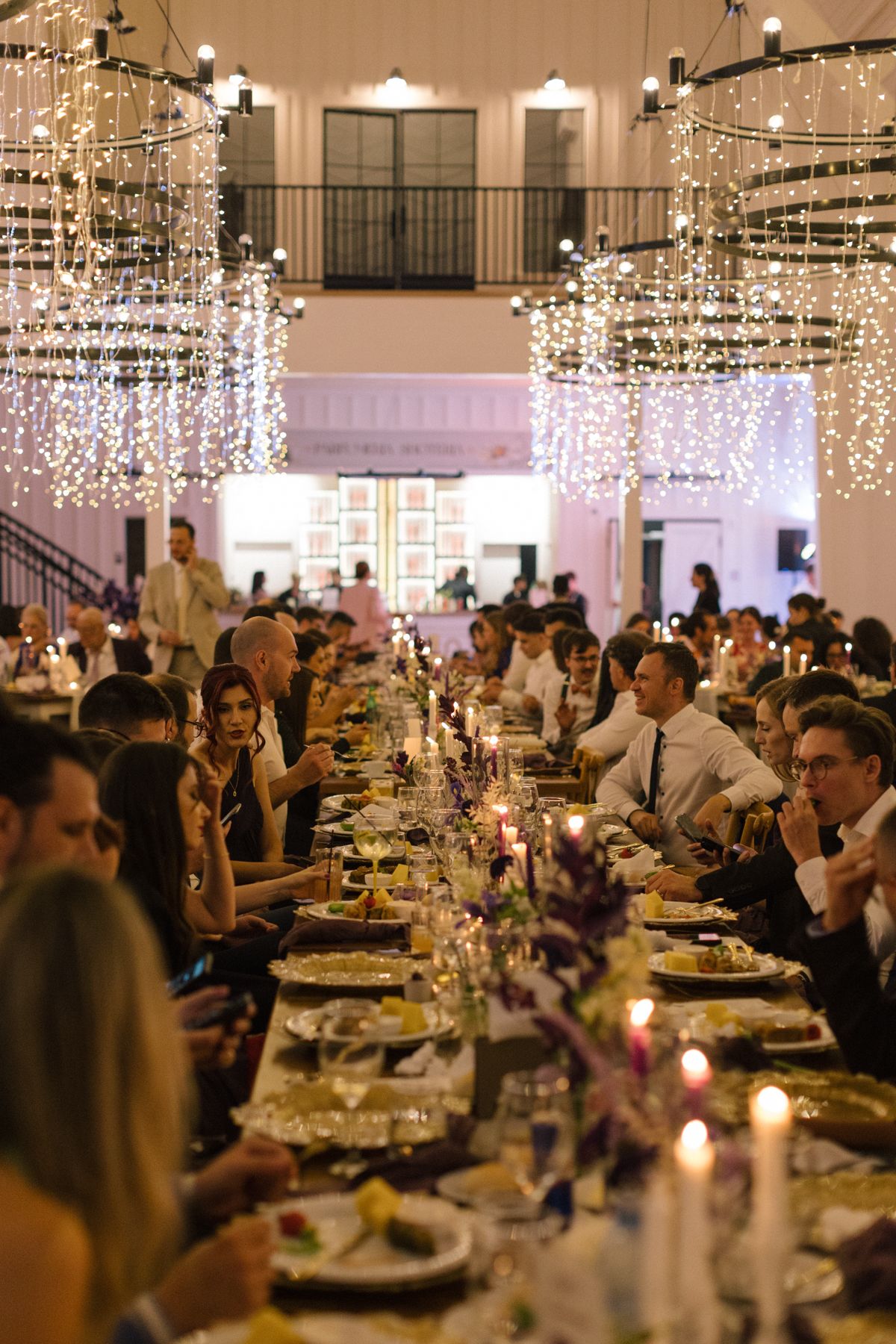 Wedding reception dinner setup with hanging crystal lights and candlelit tables in an elegant barn venue.