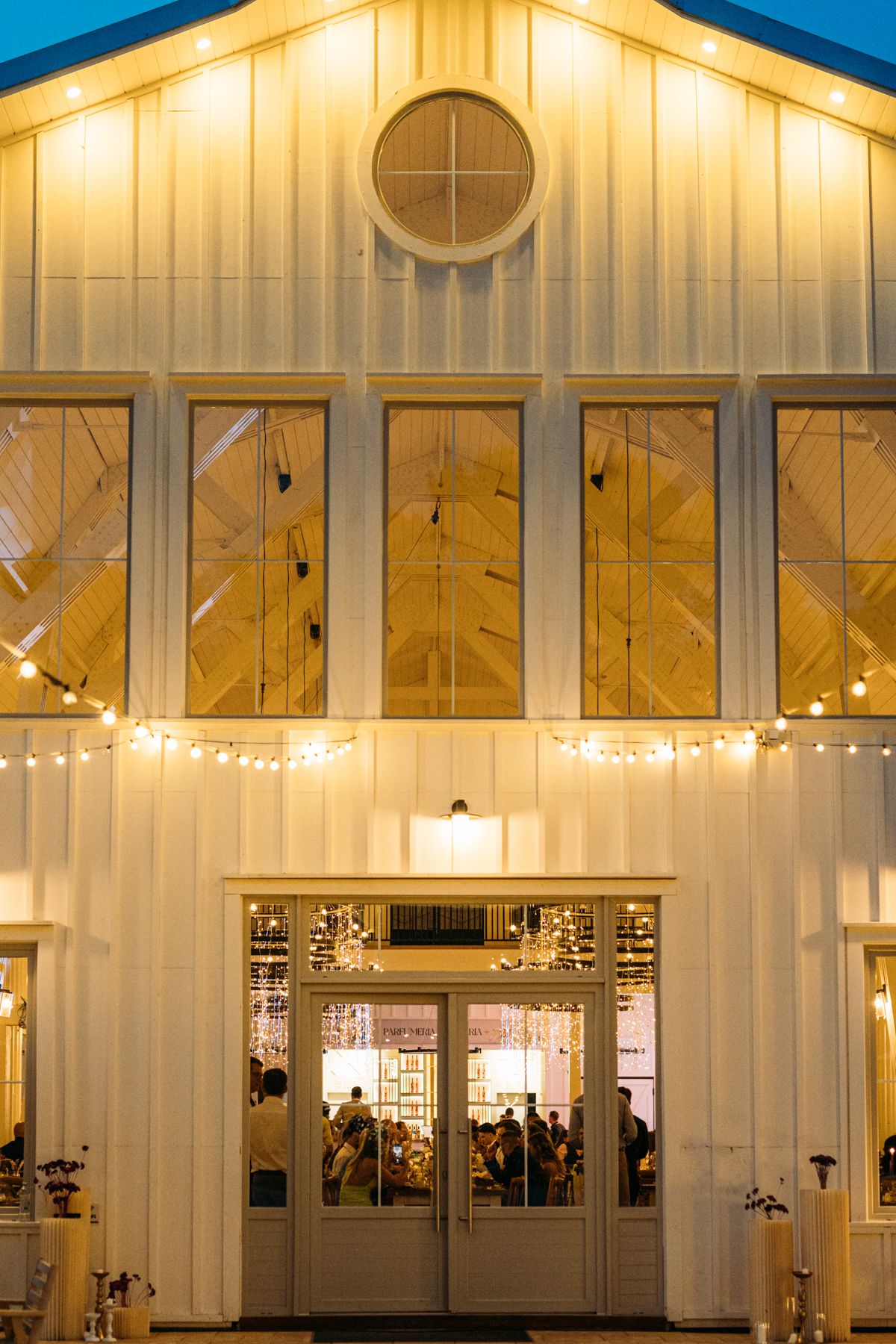 Modern white barn wedding venue facade illuminated against blue evening sky with decorative lighting.