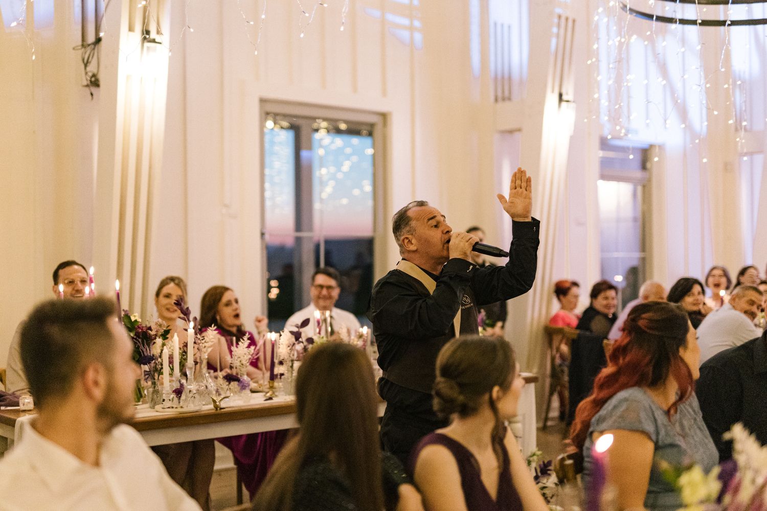 Wedding reception guests celebrating with raised arms in an elegant white-decorated venue with string lights.