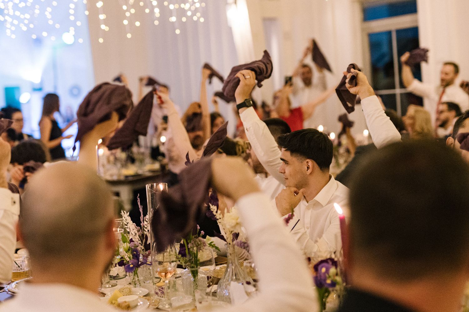 Wedding celebration with guests raising hands in a brightly lit reception hall.