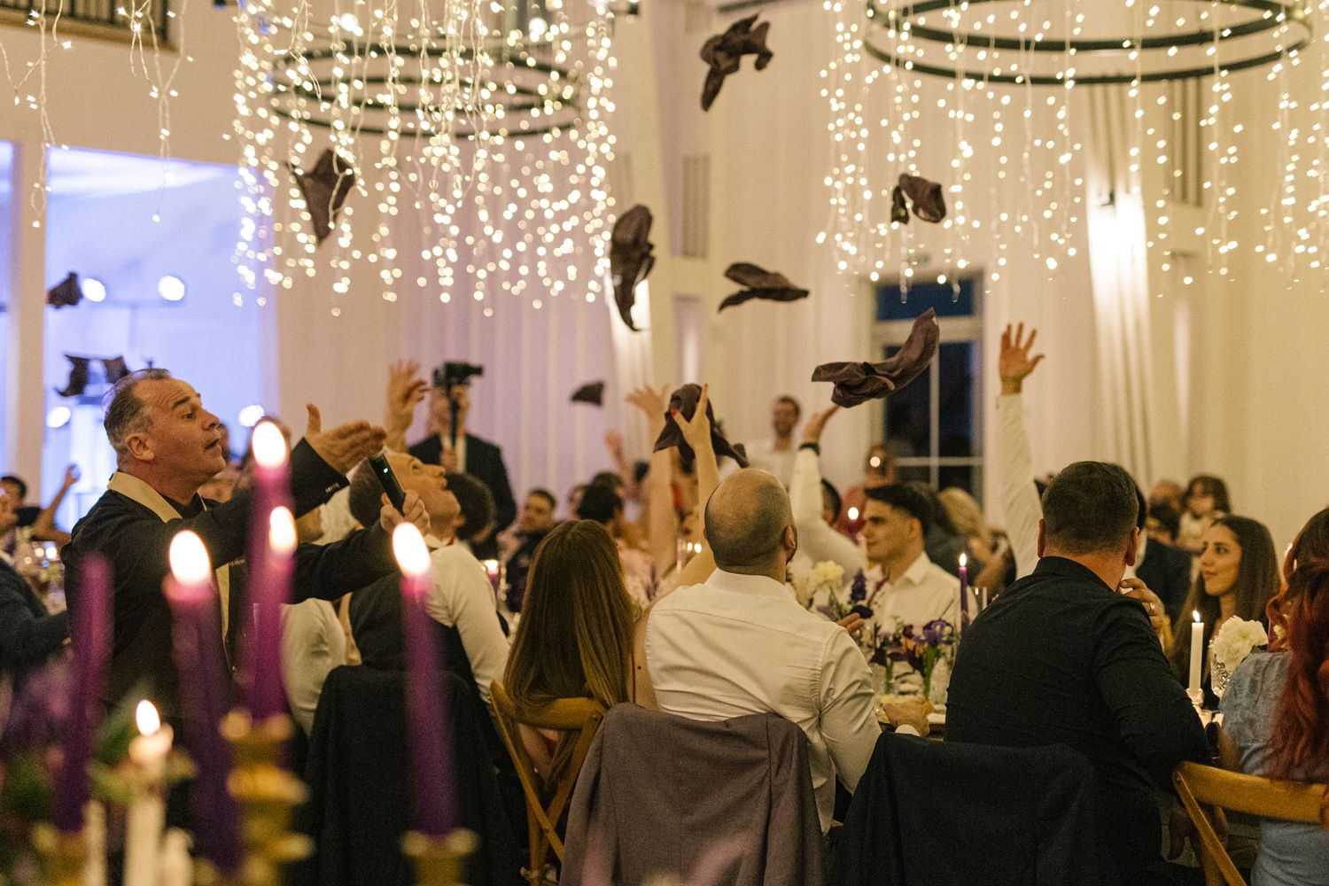 Wedding reception guests dancing under elegant crystal chandeliers and string lights at an indoor venue.