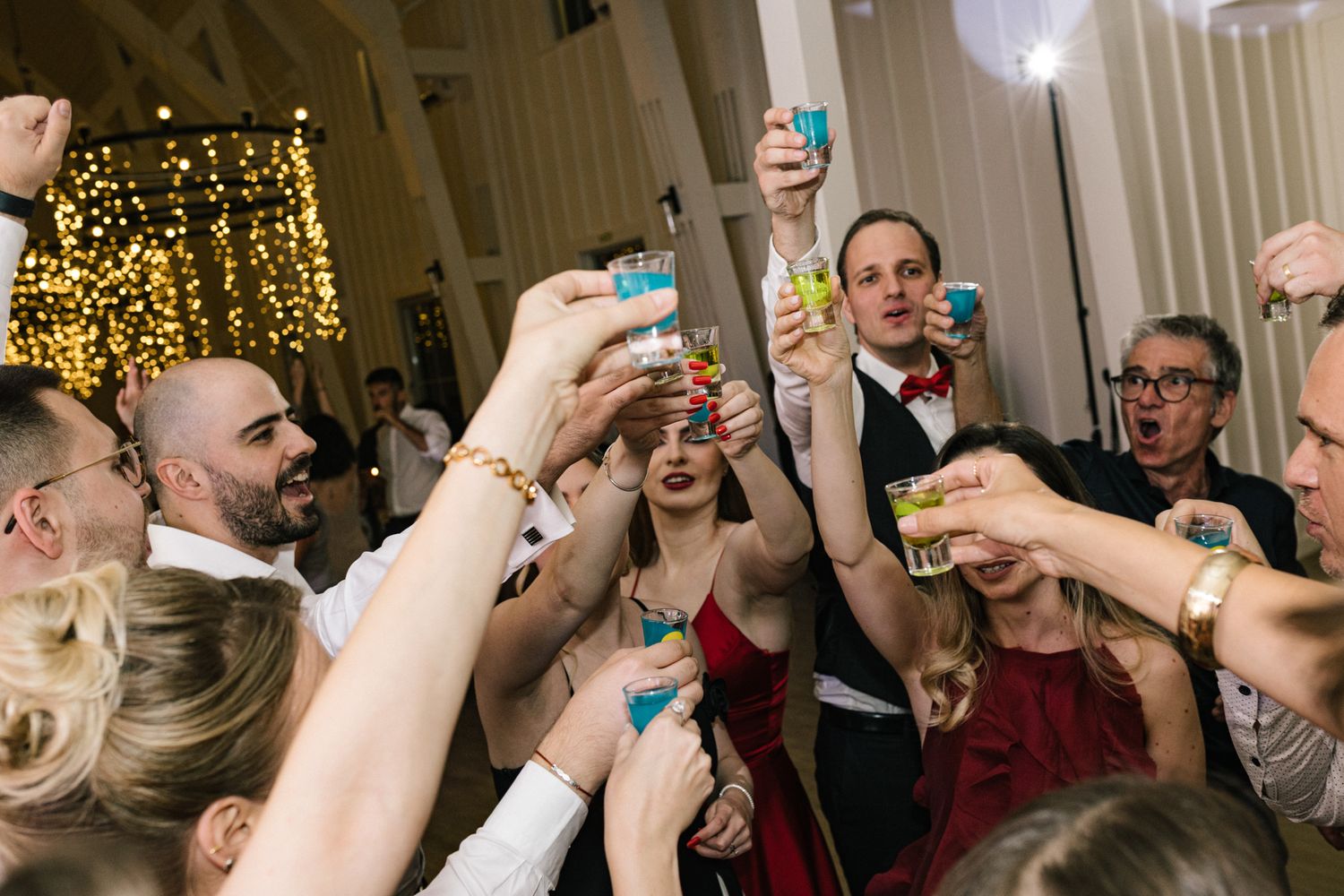Crowd raising blue shot glasses in a celebratory toast at an indoor party.