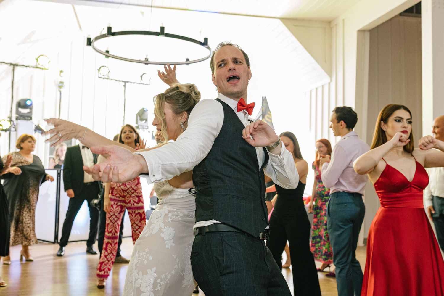 Wedding guests dancing energetically in a bright indoor venue with circular lighting fixtures overhead.