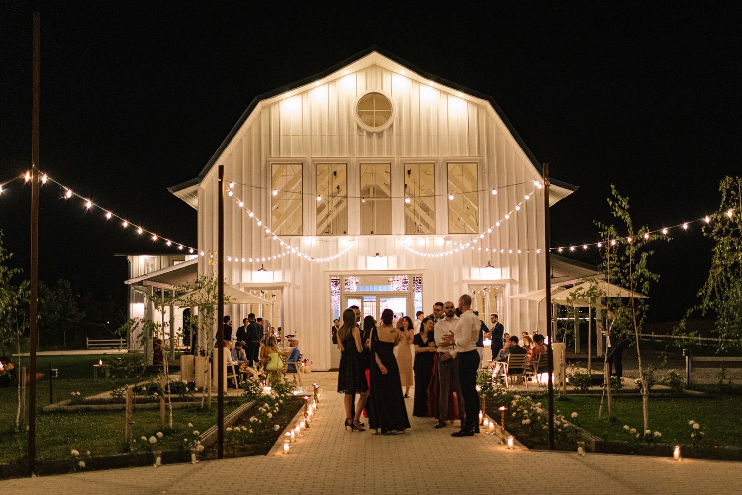 Beautiful white barn venue illuminated at night with string lights and guests gathering outside.