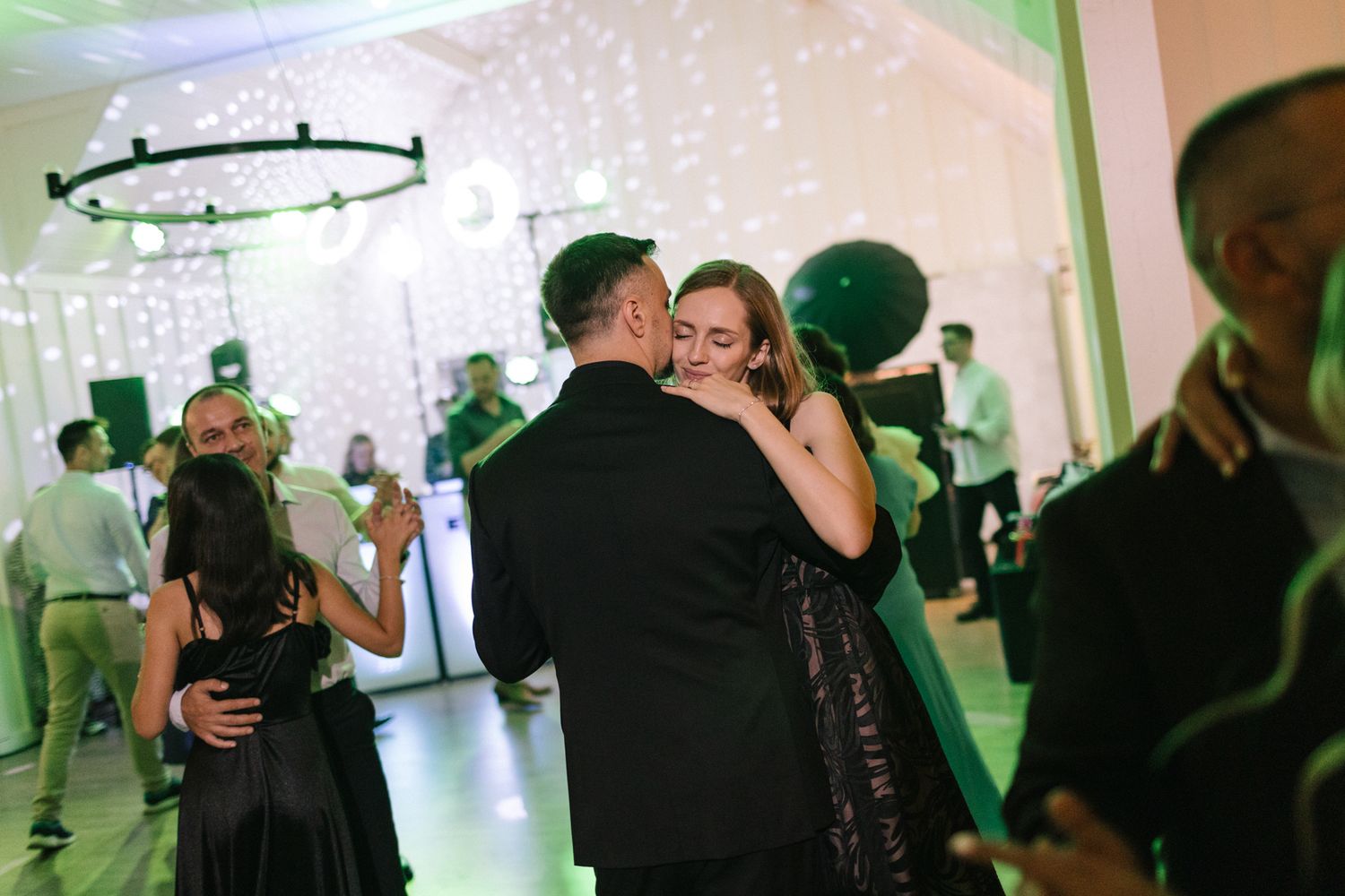 Wedding guests dance and celebrate at an indoor reception with green lighting accents.
