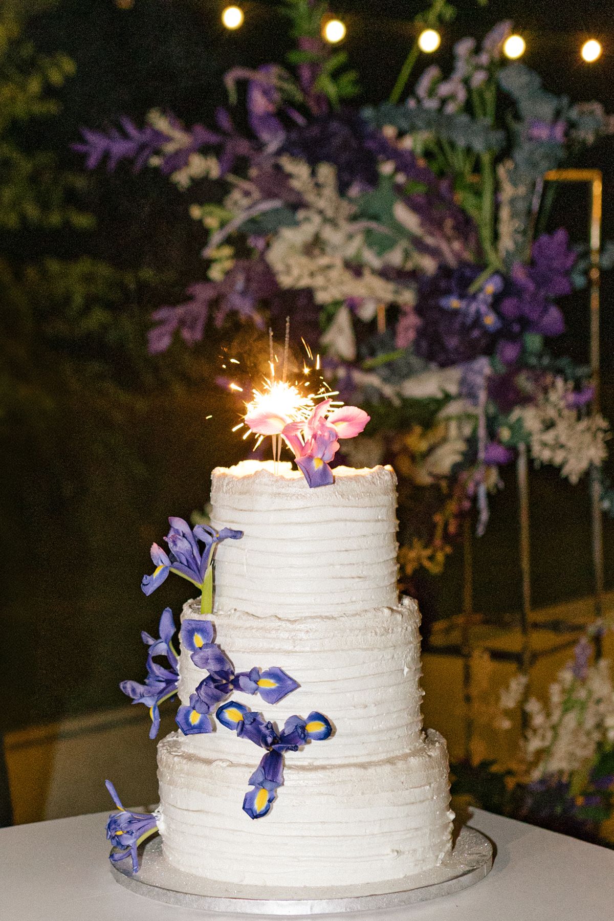 White tiered wedding cake decorated with purple iris flowers and sparklers on top.