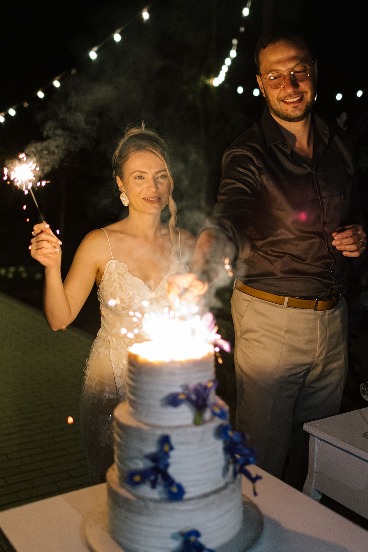 Wedding cake with sparklers and blue flowers being lit at night outdoors.