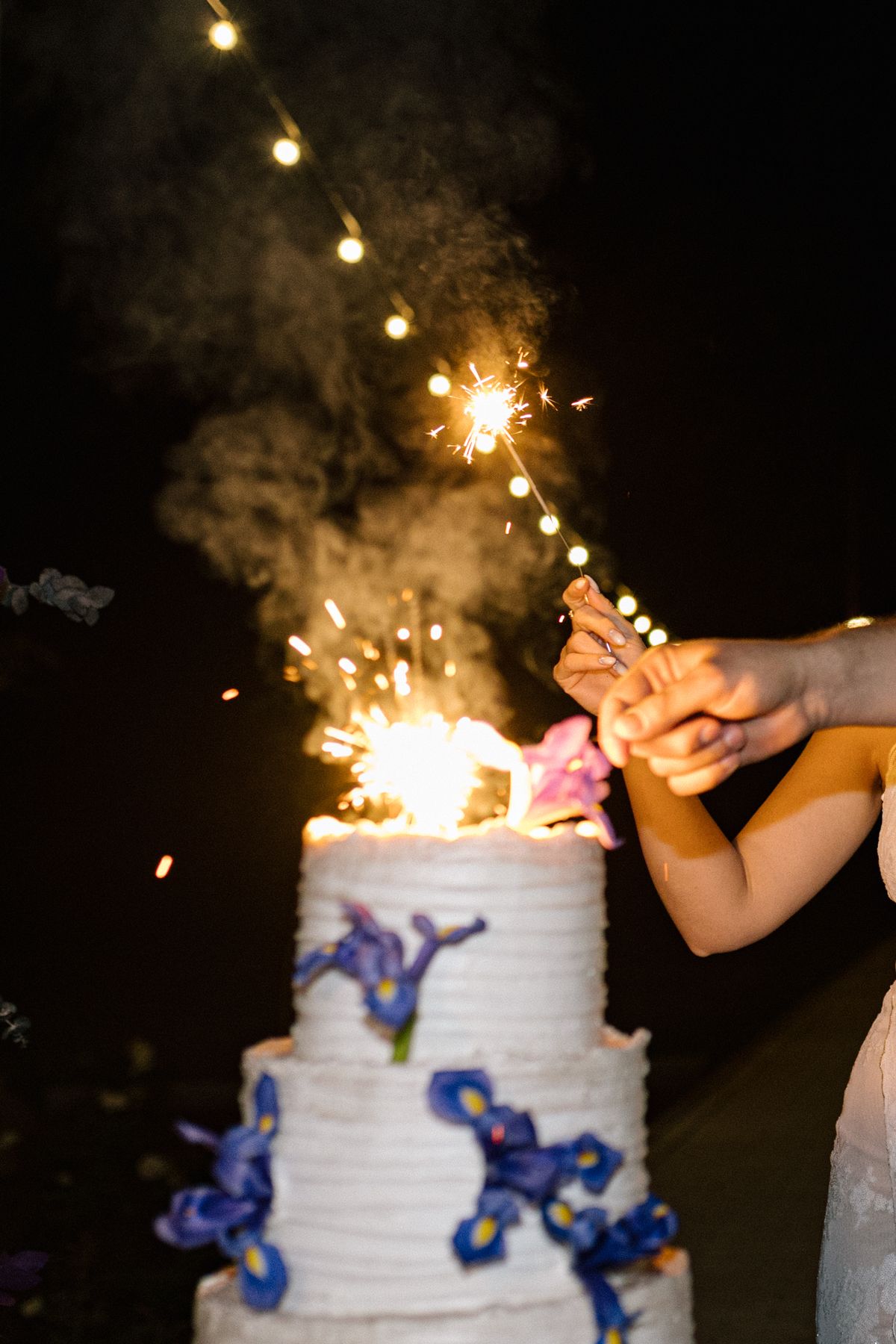 Sparklers being lit on a white wedding cake with blue floral decorations at night.