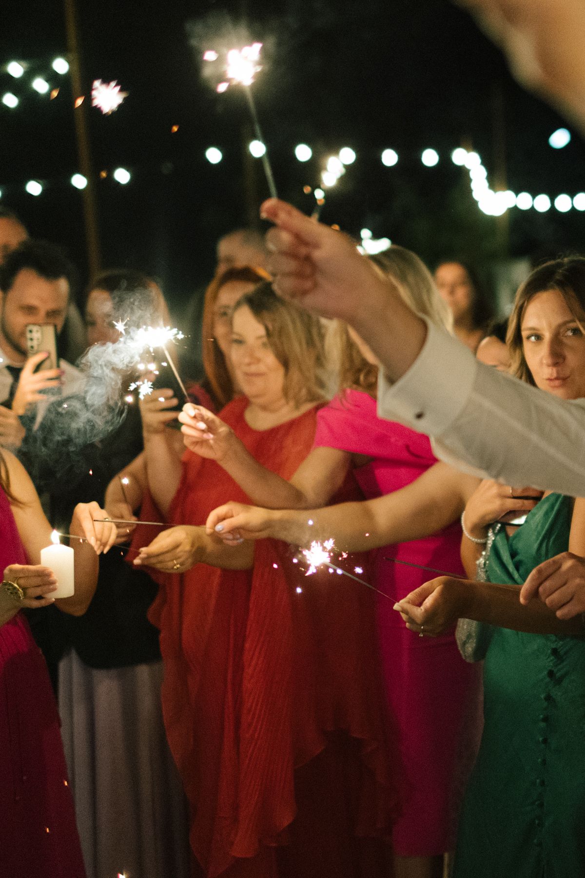 Guests in colorful dresses hold sparklers during an outdoor evening celebration.