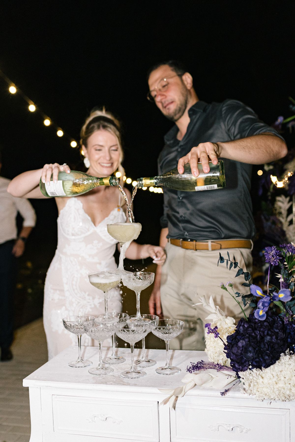 Wedding couple pours champagne into vintage coupe glasses at an elegant reception bar setup.