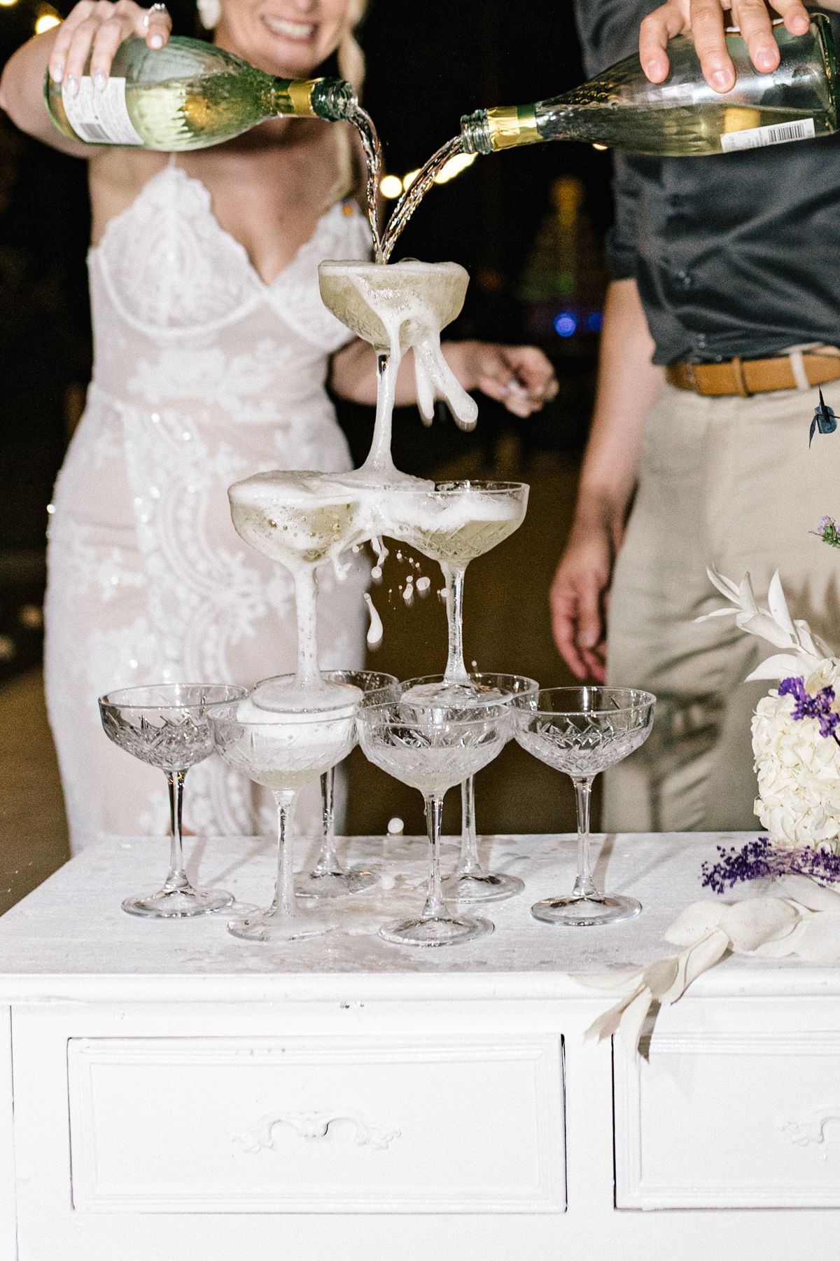 Champagne being poured into crystal coupe glasses on white table with purple floral arrangements.