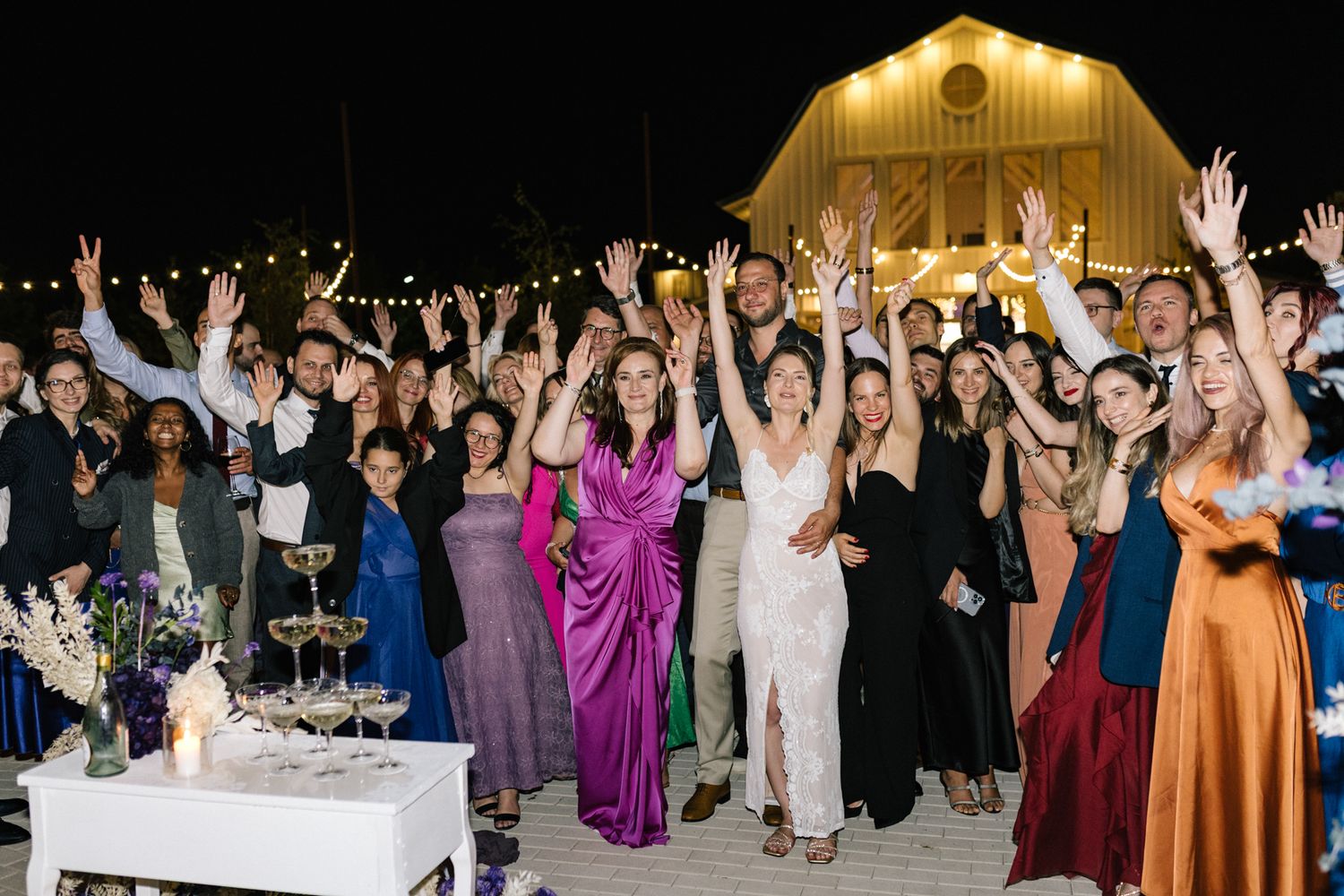 Large crowd of wedding guests cheering with arms raised at outdoor barn reception.