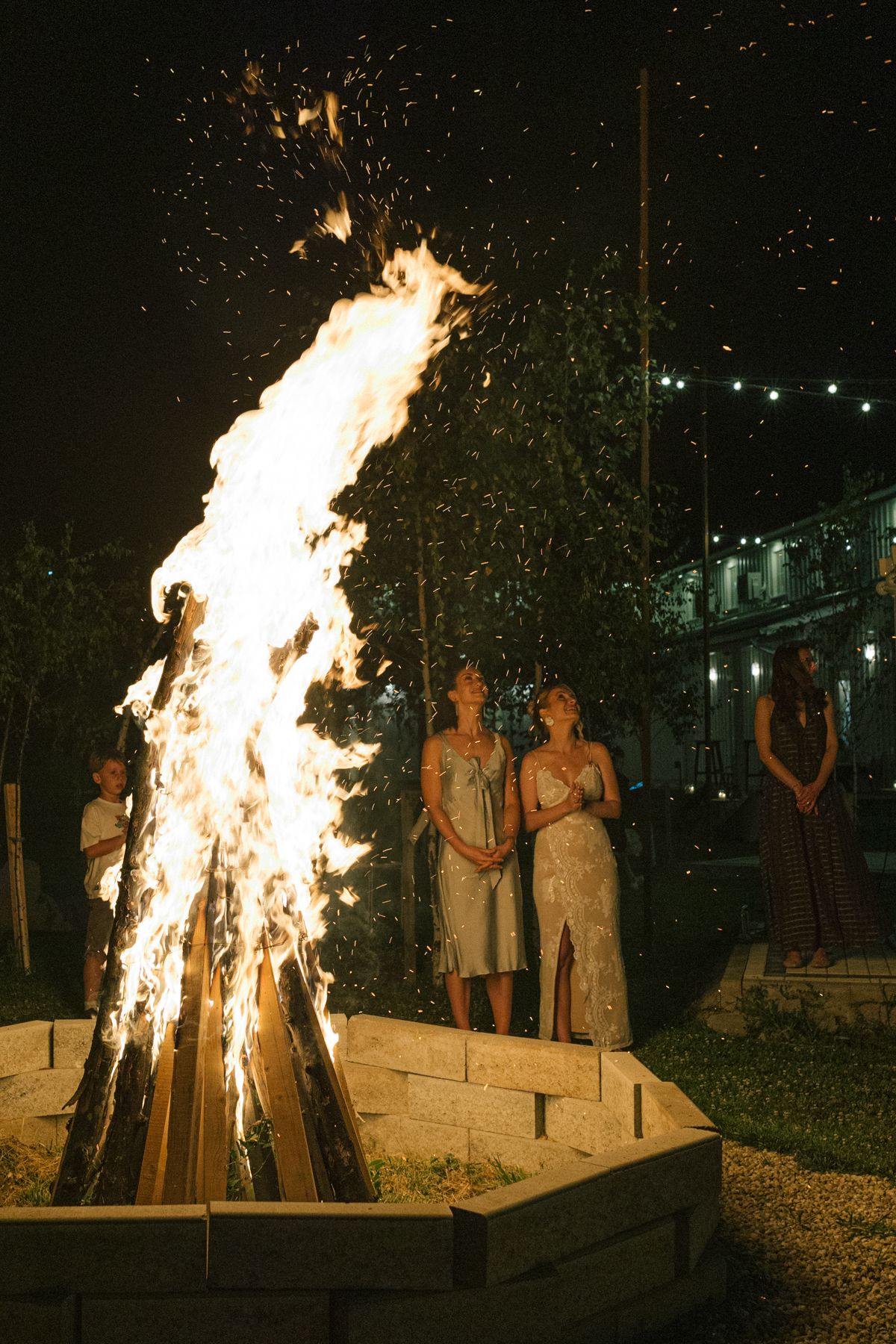 Large bonfire burning brightly at night with sparks flying as people gather around to watch the flames.