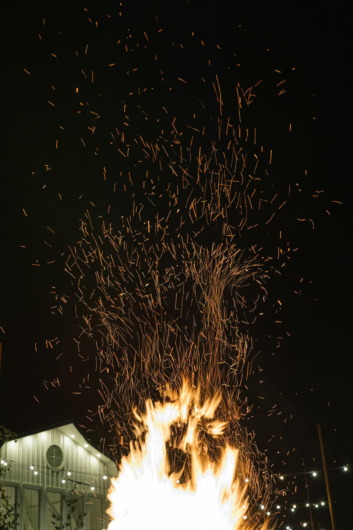 Dramatic nighttime bonfire with embers rising against dark sky and building in background.