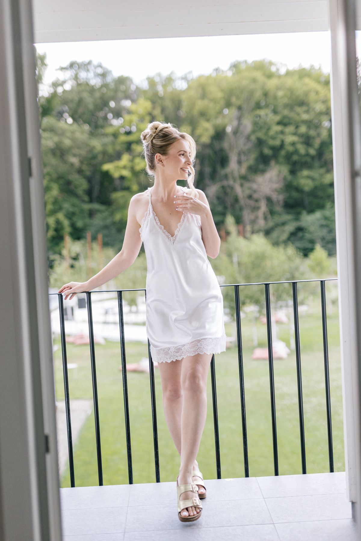 Woman in white dress poses playfully on a balcony railing with green trees in background.