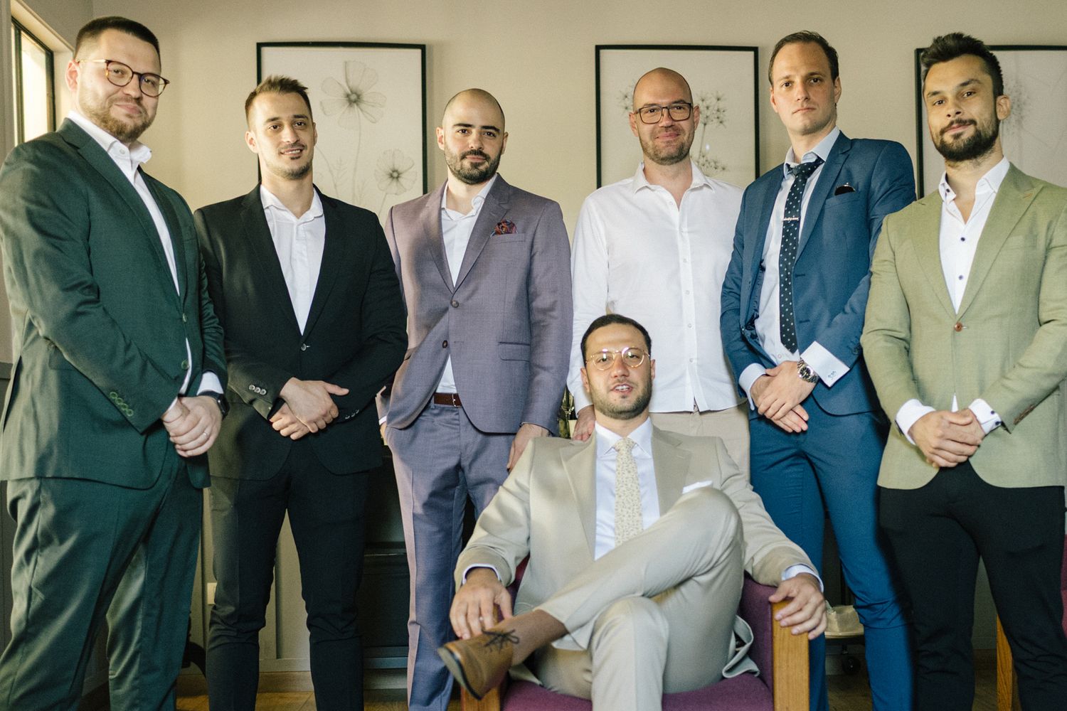 Wedding party groomsmen lined up in formal suits against a neutral wall.
