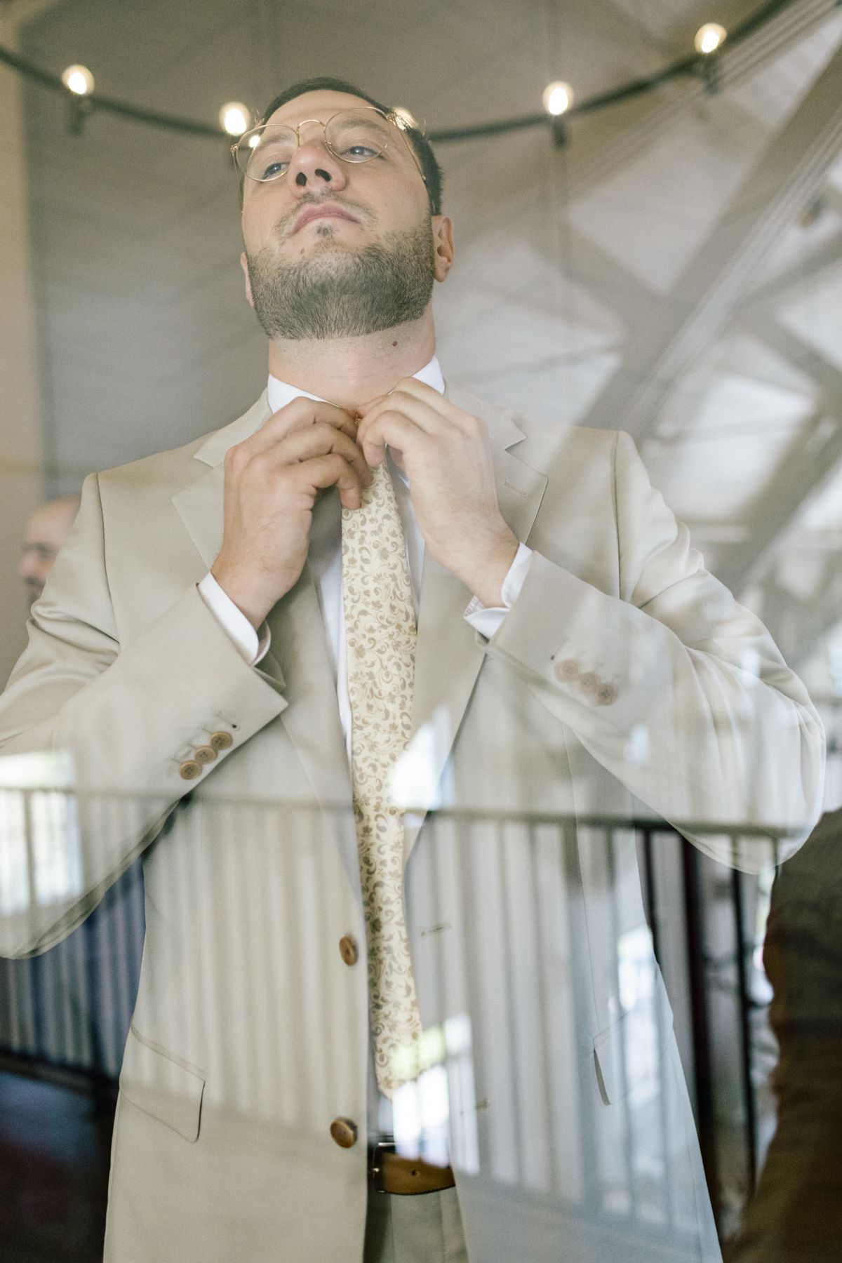 Groom adjusts his tie while wearing a light beige suit and patterned gold tie.