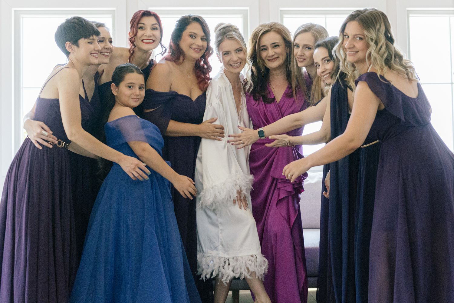 Bridal party in purple and navy dresses gather around bride in white gown for celebratory photo.