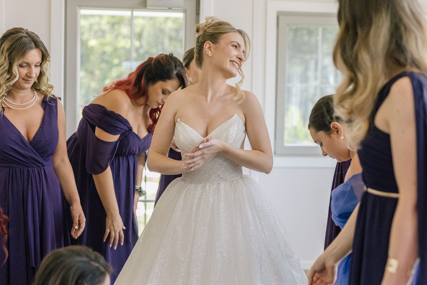 Bridesmaids in navy dresses help adjust bride's white ballgown during wedding preparations.