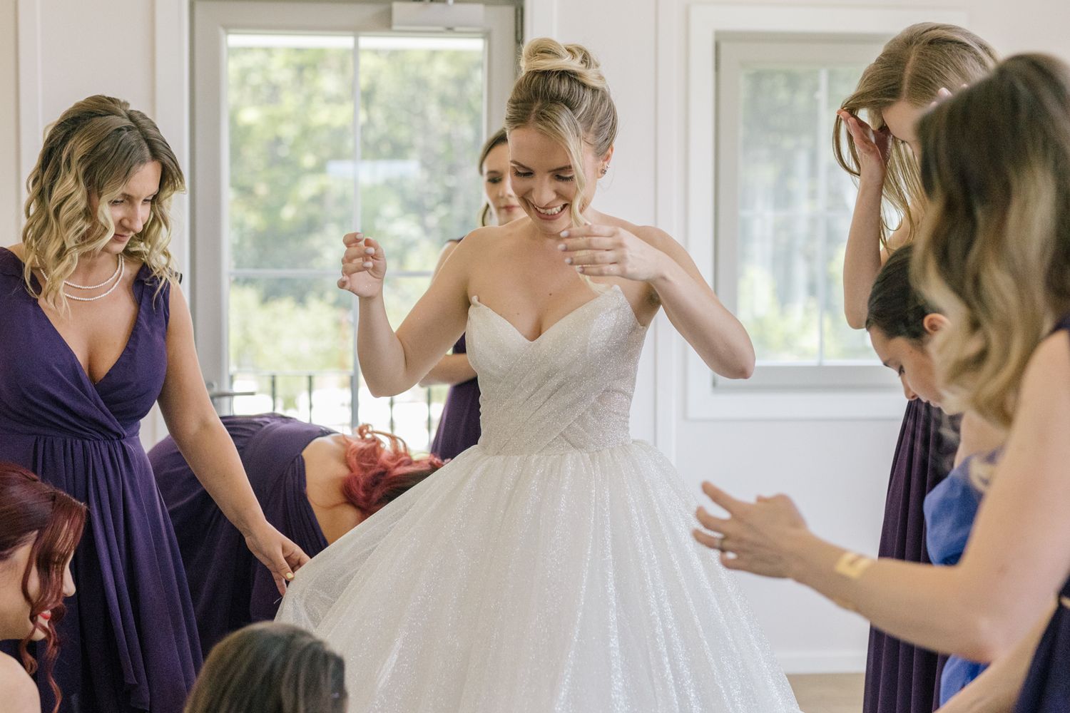 Bridesmaids in navy dresses assist bride with her white ballgown in sunlit room.