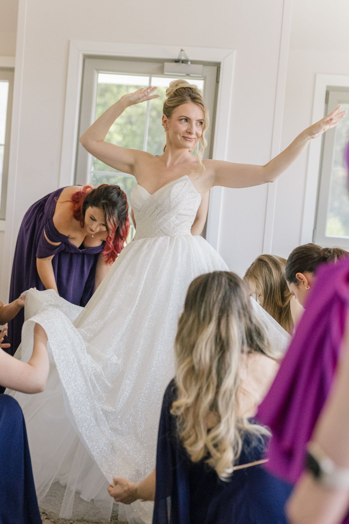 Bride in white ballgown raises arms while surrounded by bridesmaids helping with dress.