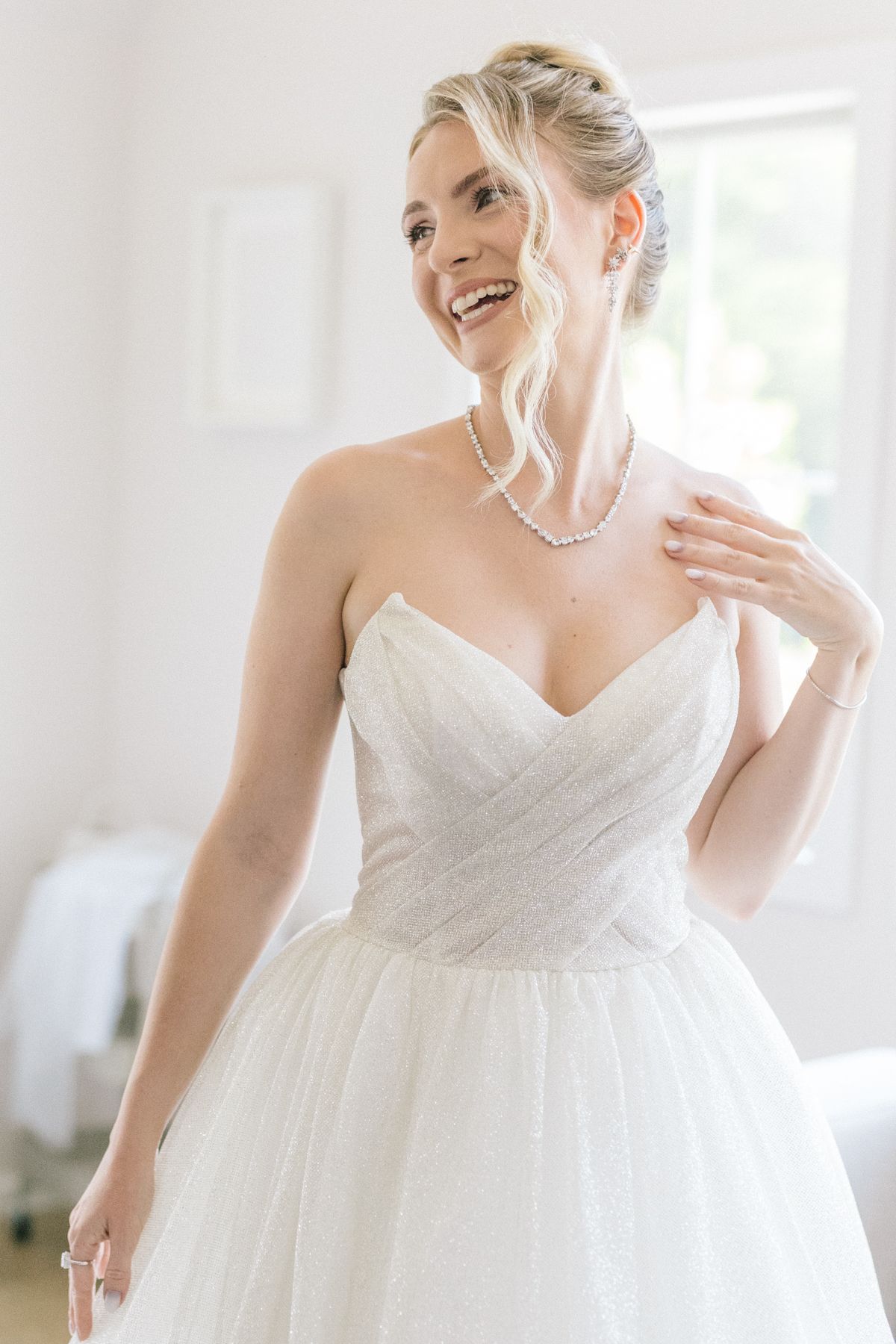 A bride in a white strapless wedding gown laughs joyfully in a bright sunlit room.