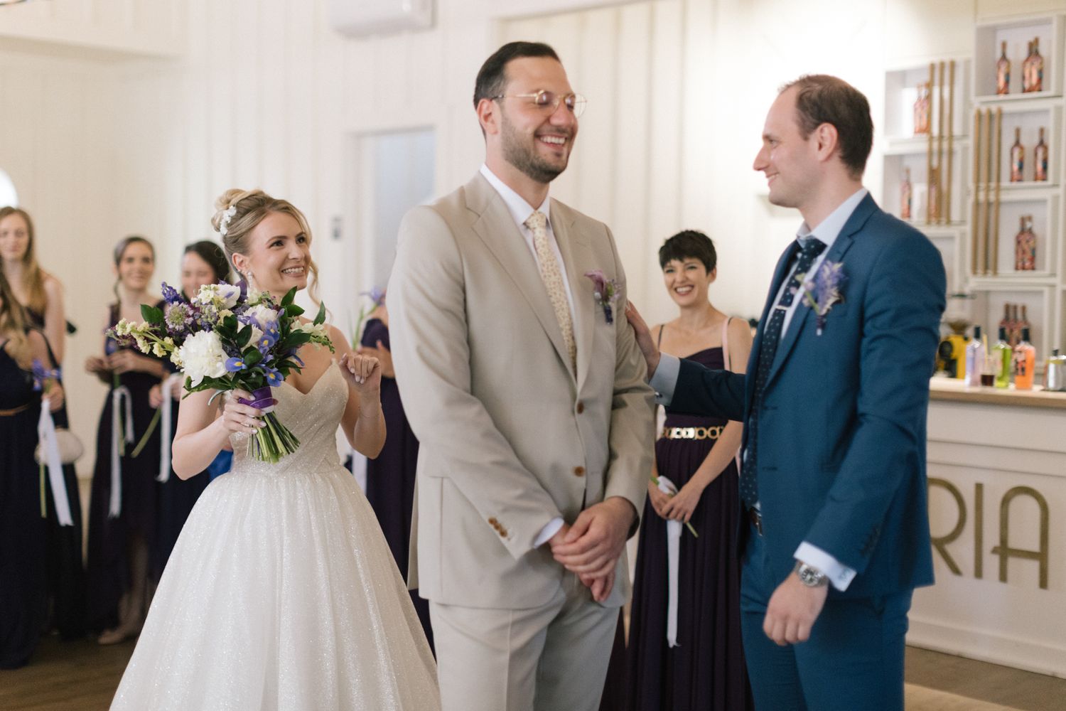 Wedding celebration in an elegant white room with guests in navy and beige formal attire.