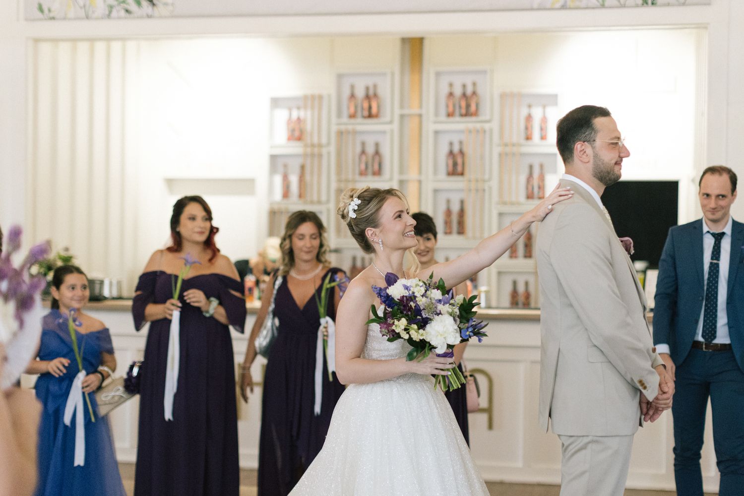 Wedding party members gather in a bright venue with white walls and decorative frames.
