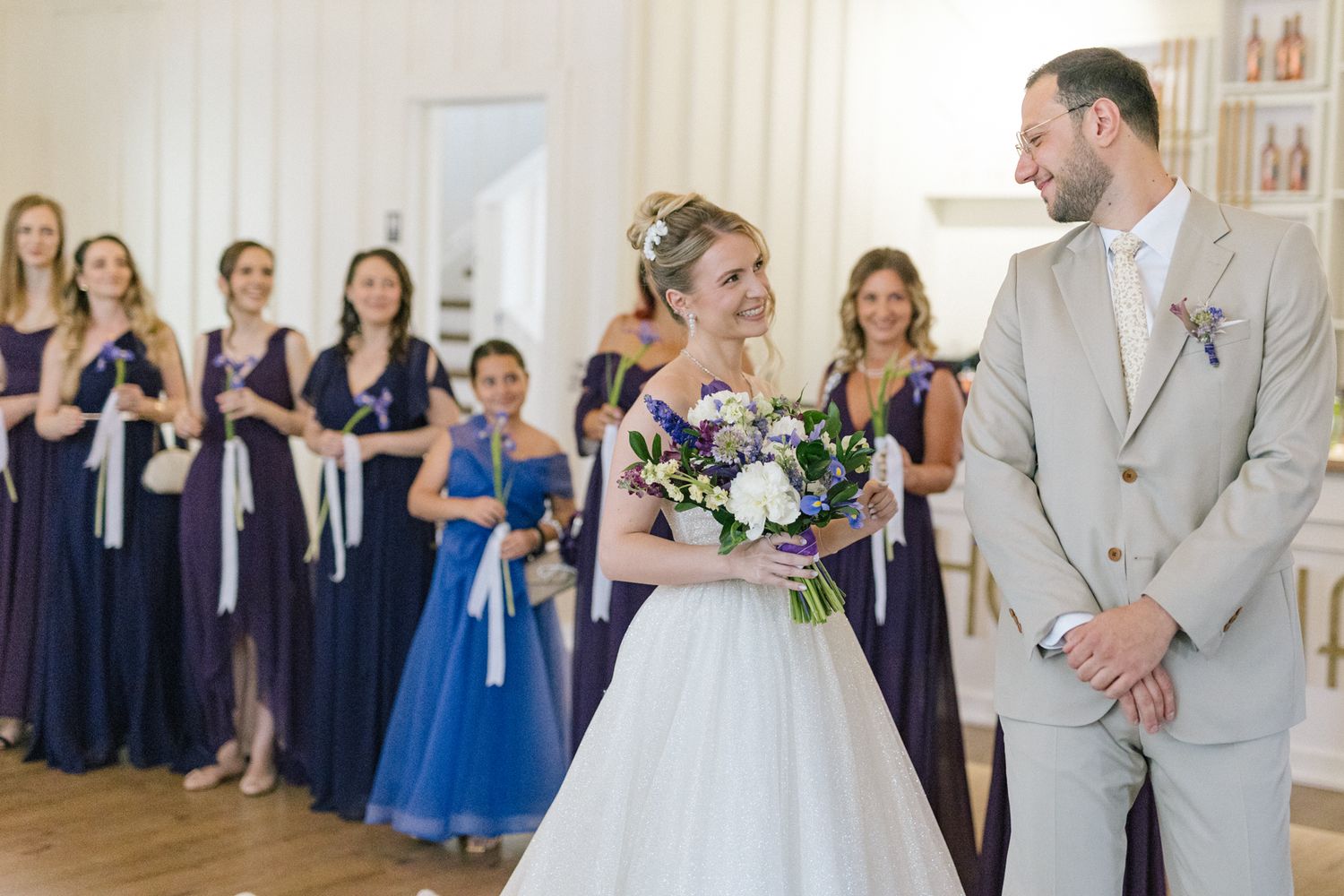 Wedding celebration scene with guests in navy dresses and formal attire in an elegant white room.