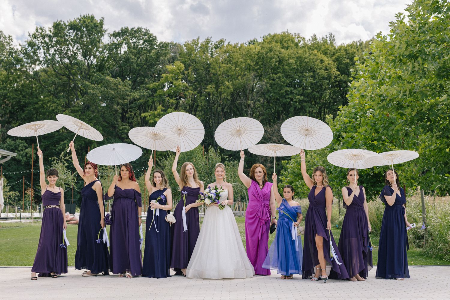 Wedding party with bridesmaids in purple and navy dresses posing outdoors with white parasols.