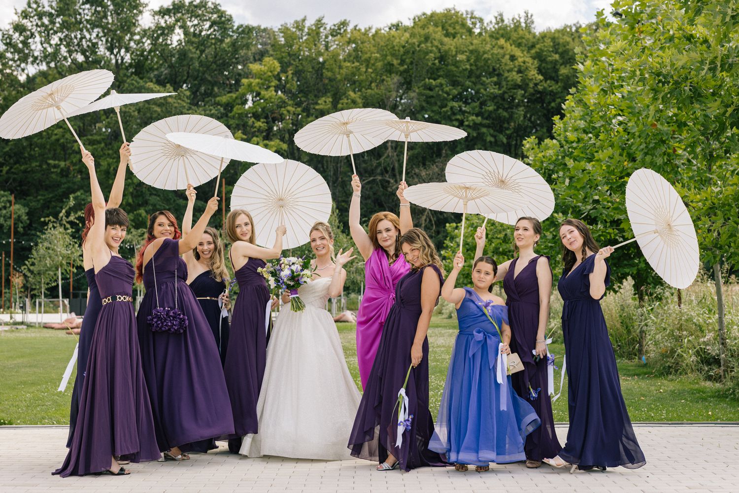 Joyful bridal party celebrating with white parasols in outdoor garden setting.
