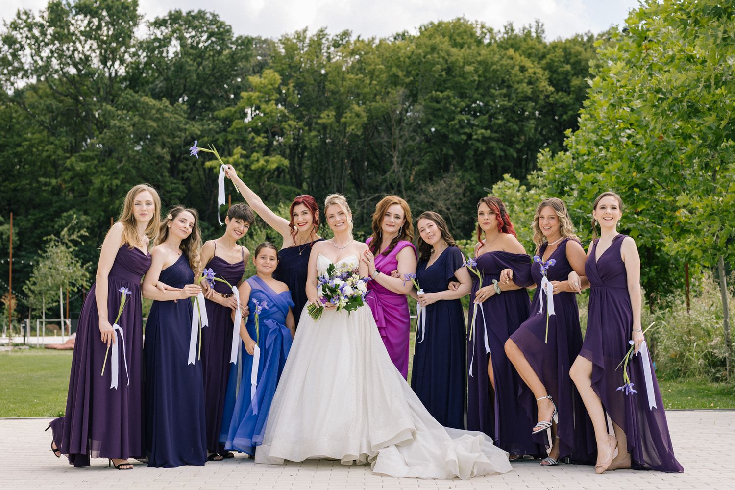 Wedding party lineup with bridesmaids in purple and navy dresses against green landscape.